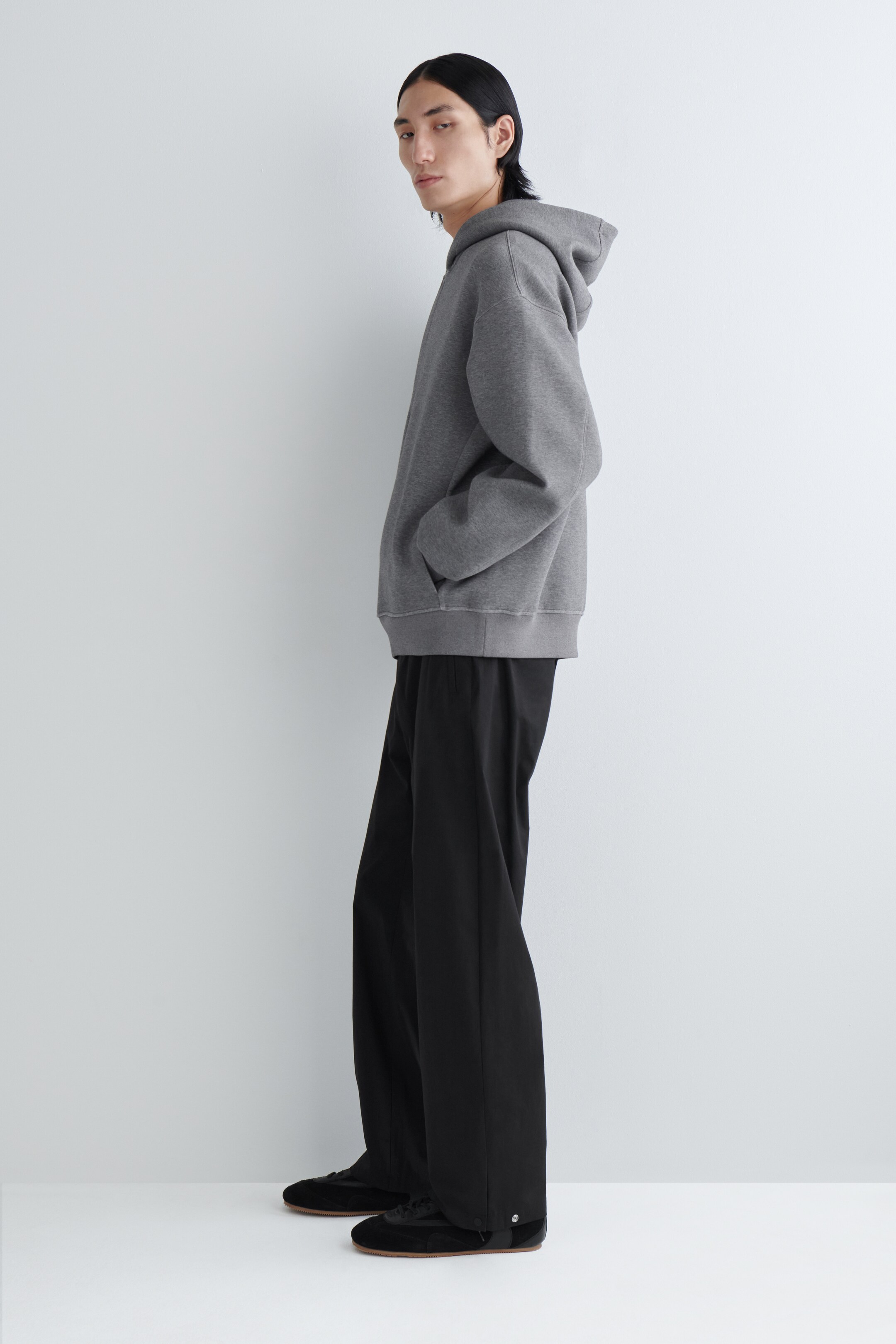 Zobrazit větší obrázek: Šedá mikina s kapucí a černé kalhoty jsou předváděny osobou stojící z profilu, čelem doprava. Šedá mikina s kapucí v uvolněném střihu je navržena s kapucí, žebrovanými manžetami, žebrovaným lemem a přední kapsou typu klokanka, a černé kalhoty s širokými nohavicemi se objevují s detaily na patentky u pravého kotníku.