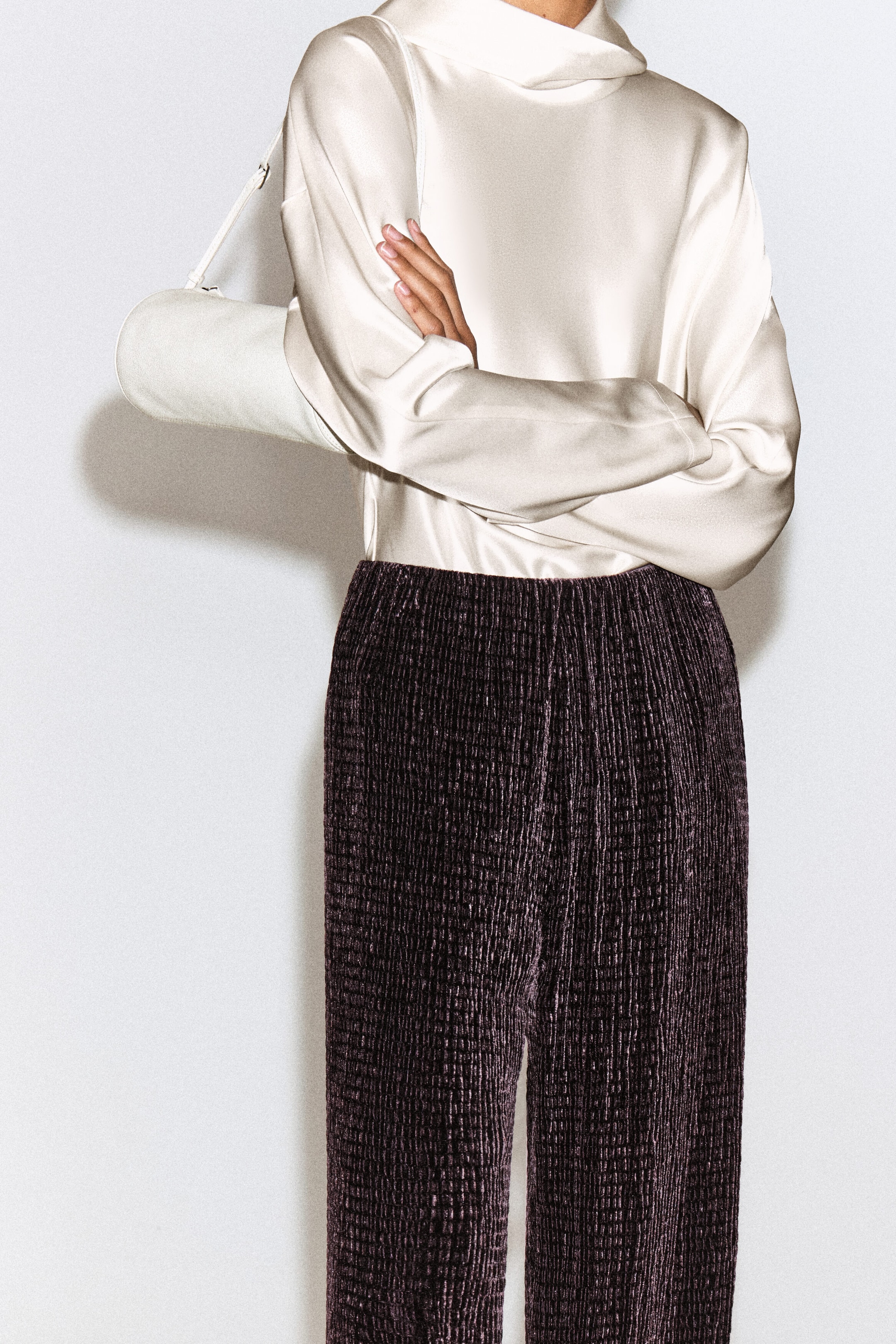 A cream-colored, long-sleeved satin blouse featuring a draped cowl neck is shown tucked into dark purple, textured ribbed velvet trousers with a wide-leg silhouette, facing front. A white cylindrical shoulder bag is styled under the crossed arm to complement the outfit. A cream-colored, long-sleeved satin blouse featuring a draped cowl neck is shown tucked into dark purple, textured ribbed velvet trousers with a wide-leg silhouette, facing front. A white cylindrical shoulder bag is styled under the crossed arm to complement the outfit.