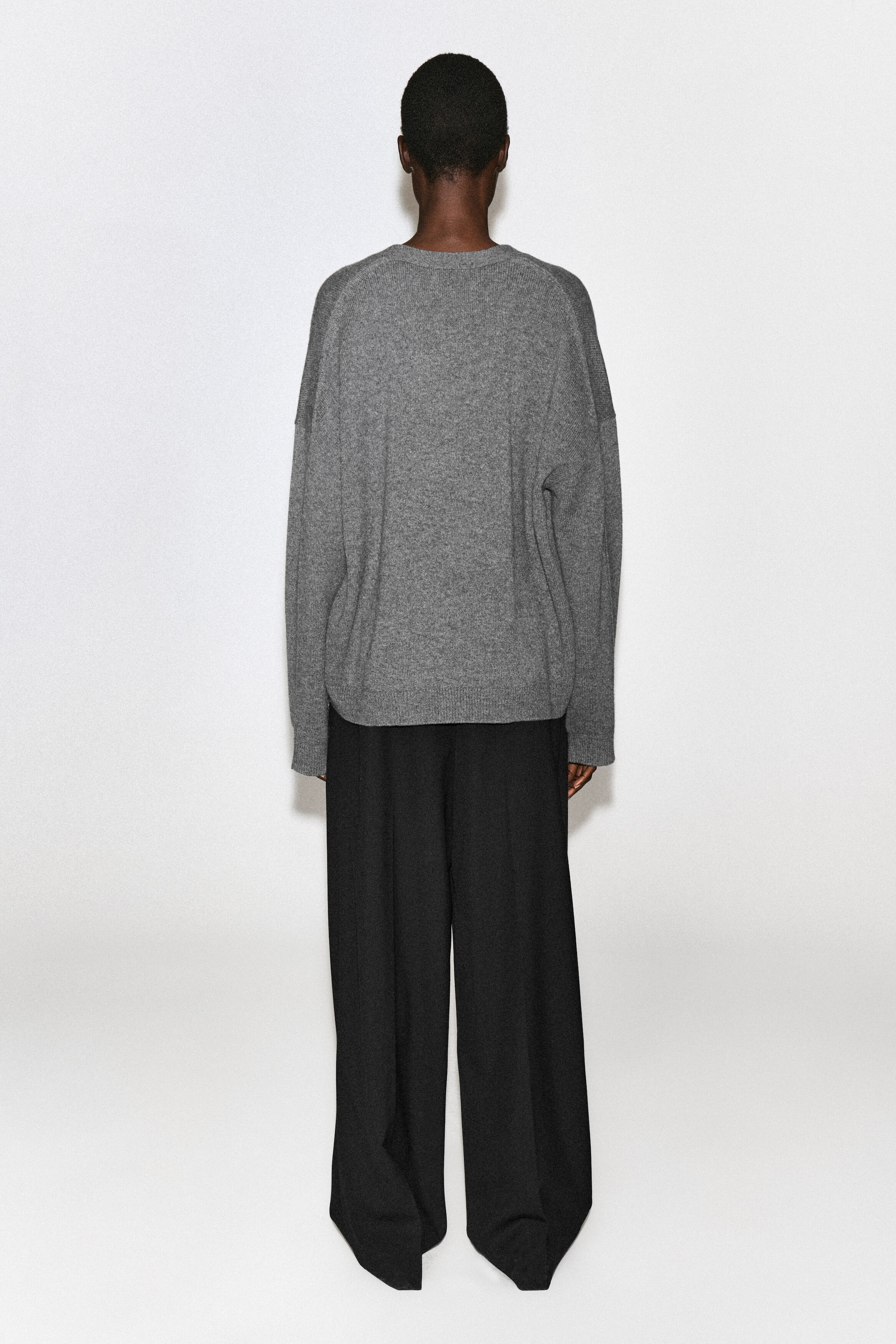 Una modella indossa un cardigan in cashmere grigio, mostrato da dietro con una vestibilità rilassata, spalle scese e fondo a costine. Il cardigan in maglia è abbinato a pantaloni neri a gamba larga. Una modella indossa un cardigan in cashmere grigio, mostrato da dietro con una vestibilità rilassata, spalle scese e fondo a costine. Il cardigan in maglia è abbinato a pantaloni neri a gamba larga.