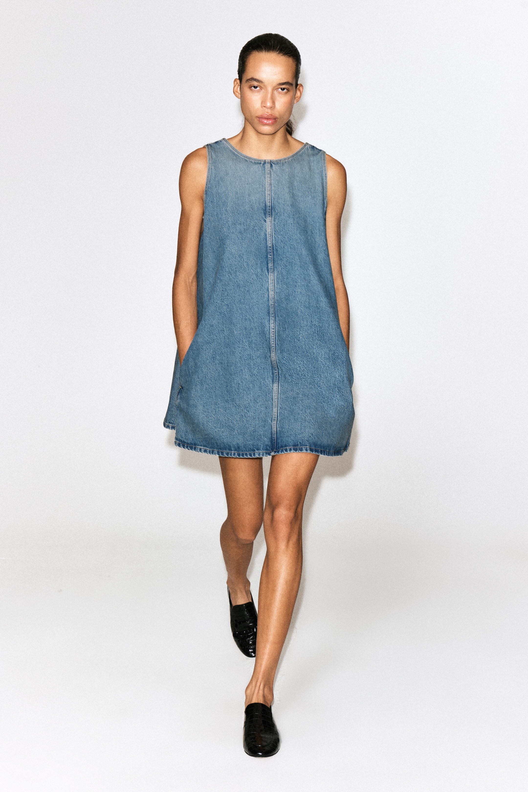 Un mini abito in denim senza maniche, in lavaggio blu medio, è mostrato frontalmente, mettendo in risalto una silhouette ad A con girocollo e una prominente cucitura verticale centrale. L’abito è dotato di tasche laterali ed è abbinato a mocassini in pelle nera.