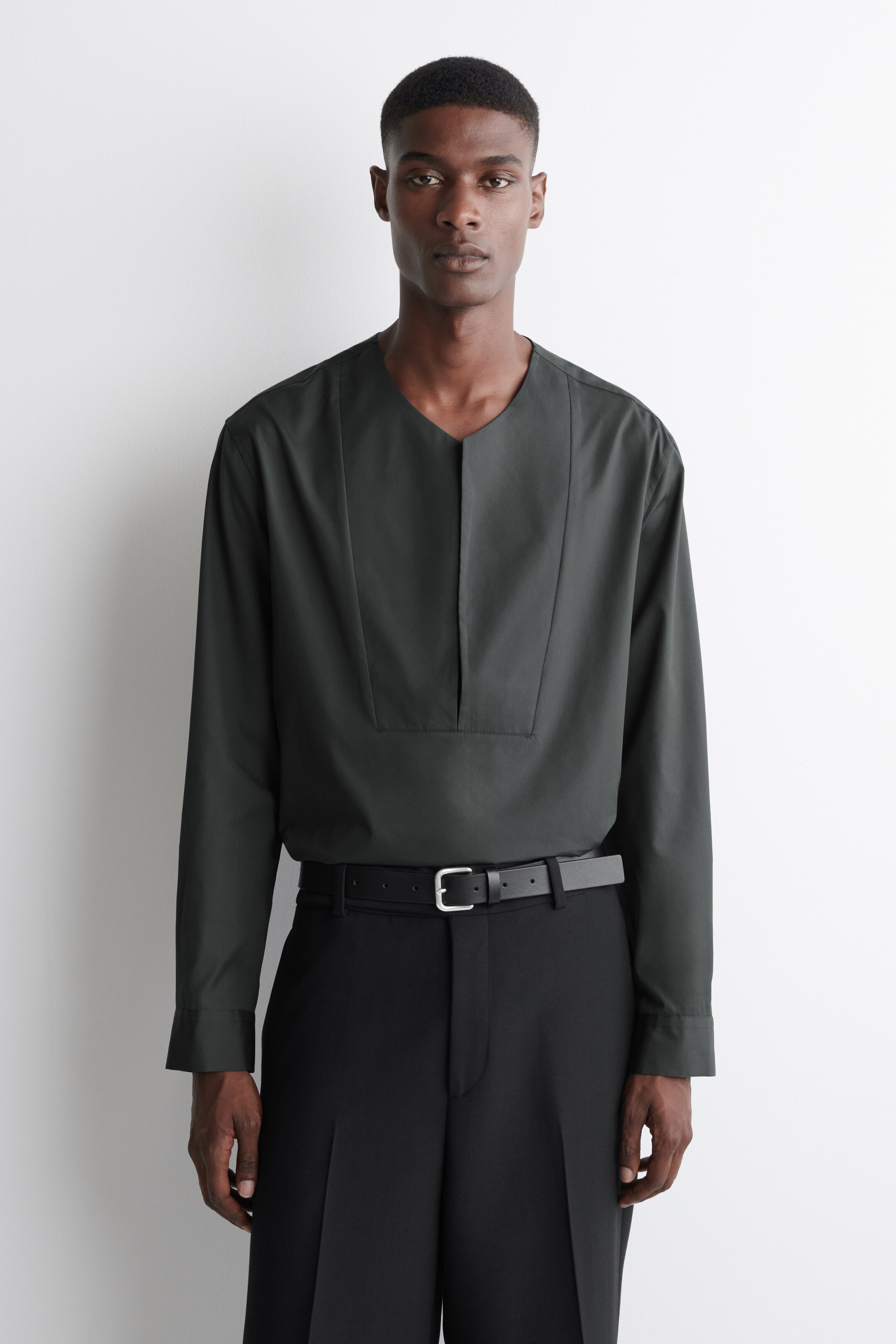 Ver imagen más grande: Una camisa de manga larga de color verde oscuro se lleva de frente, presentando un escote en V dividido y dos paneles verticales estructurados que se extienden desde los hombros hasta una costura horizontal a través del pecho. La camisa se mete dentro de unos pantalones negros, con un cinturón negro visible.