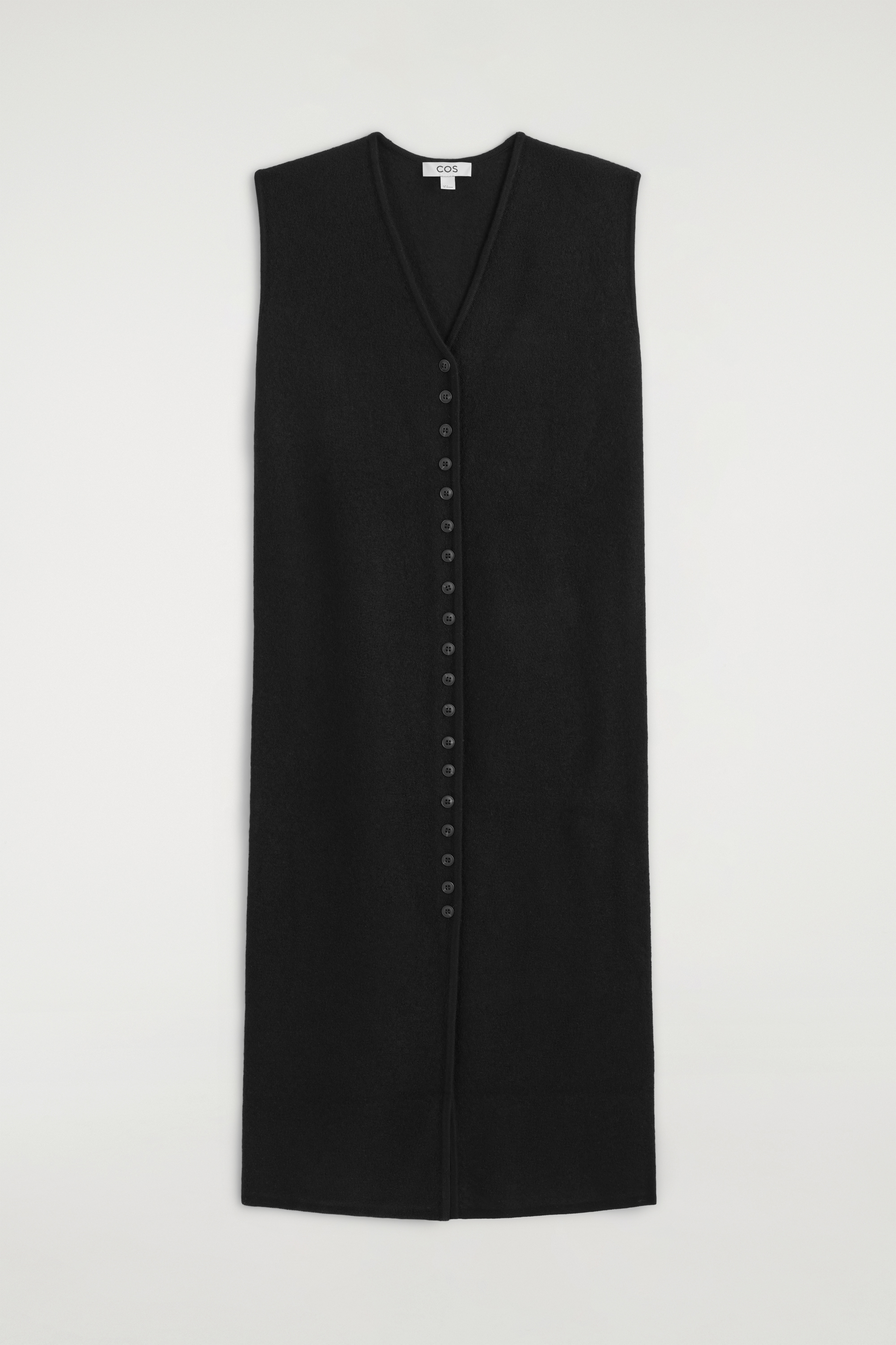The model is wearing a black boiled merino wool dress. The sleeveless midi dress features a V-neckline, a button-through front, and a front slit, and is styled with a black leather handbag, sunglasses, and black loafers.