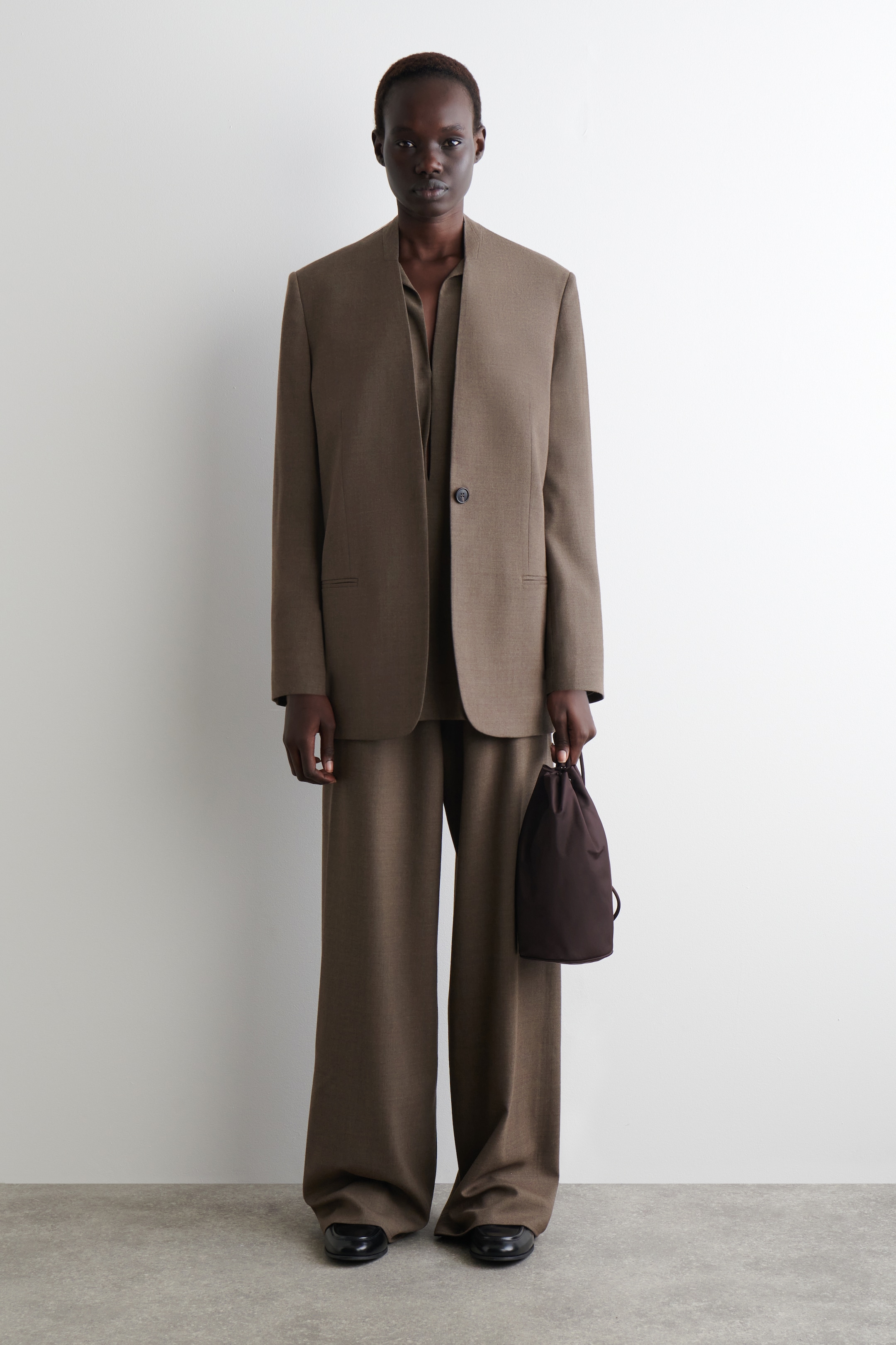View larger image: A brown suit is being modeled, facing front, consisting of a single-breasted, one-button blazer with a collarless neckline and two flap pockets, and wide-leg, full-length trousers. A matching V-neck top is being layered underneath, and a small dark brown drawstring bag is being held.