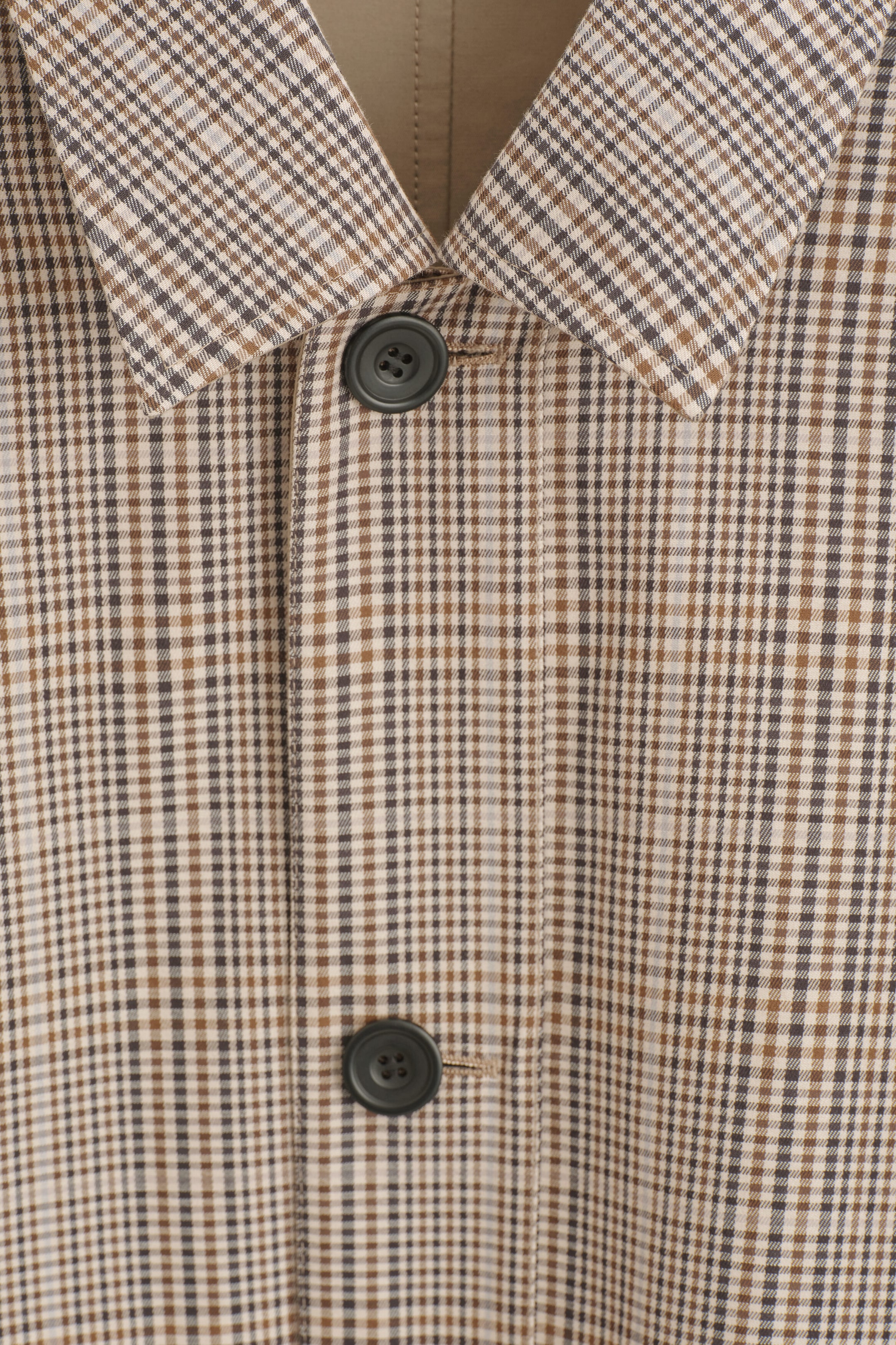 Le manteau automobile à carreaux réversible est présenté en gros plan de face, mettant en valeur son col pointu et sa patte de boutonnage avec deux boutons gris foncé. Le manteau affiche un motif à petits carreaux marron, beige et noir, avec une portion de l’envers uni beige fauve visible à l’intérieur du col. Le manteau automobile à carreaux réversible est présenté en gros plan de face, mettant en valeur son col pointu et sa patte de boutonnage avec deux boutons gris foncé. Le manteau affiche un motif à petits carreaux marron, beige et noir, avec une portion de l’envers uni beige fauve visible à l’intérieur du col.