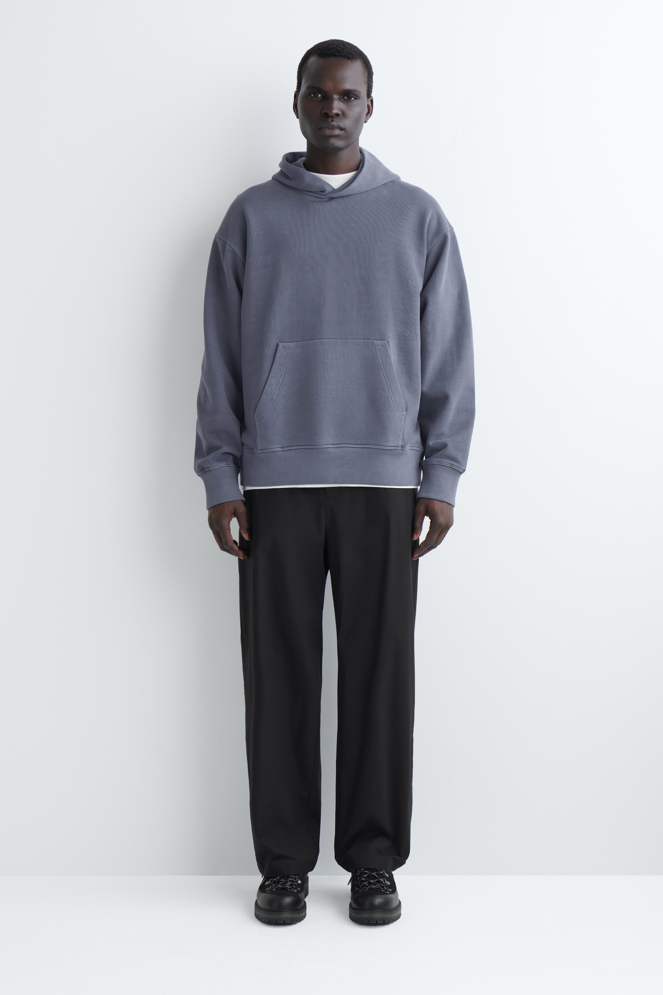 Grösseres Bild ansehen: Das Produkt ist ein nach vorne gerichteter, gedämpfter blau-grauer Kapuzenpullover mit entspannter Passform und verfügt über eine Kängurutasche, gerippte Bündchen und einen gerippten Saum. Ein weißes T-Shirt ist sichtbar darunter geschichtet, während schwarze, locker sitzende Hosen und schwarze, klobige Schnürschuhe das Outfit komplettieren.