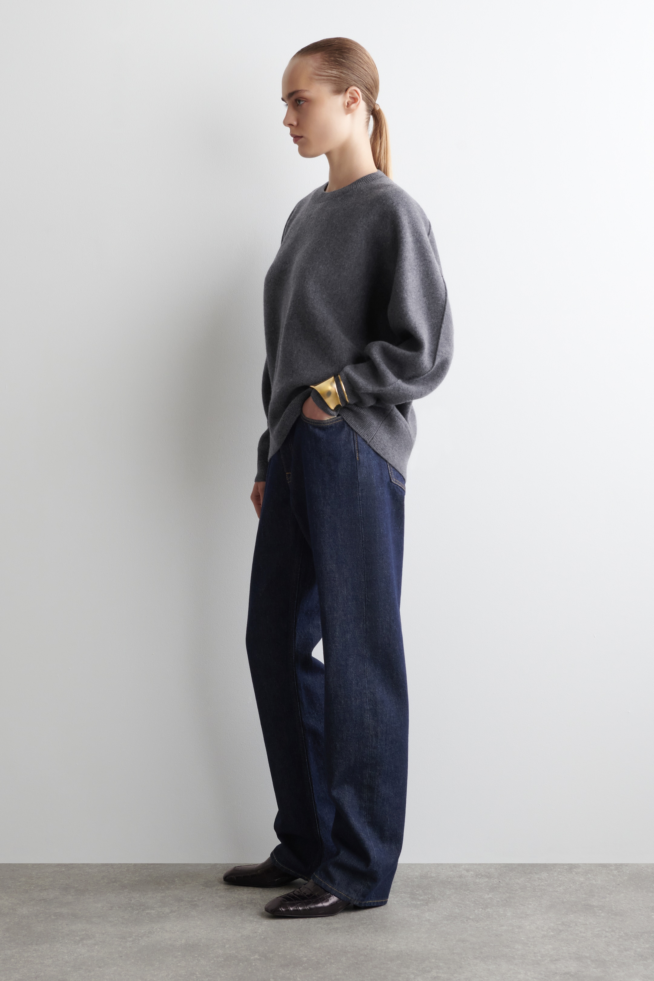Ingrandisci l'immagine: Un maglione girocollo grigio carbone mostra una vestibilità rilassata e oversize da un'angolazione laterale, con le maniche lunghe che si estendono fino ai polsi e coprono parzialmente un ampio polsino dorato. È abbinato a jeans a gamba larga indaco scuro che cadono in un drappeggio sciolto e a tutta lunghezza fino alle caviglie, dove vengono indossate scarpe basse scure e testurizzate con punte appuntite.