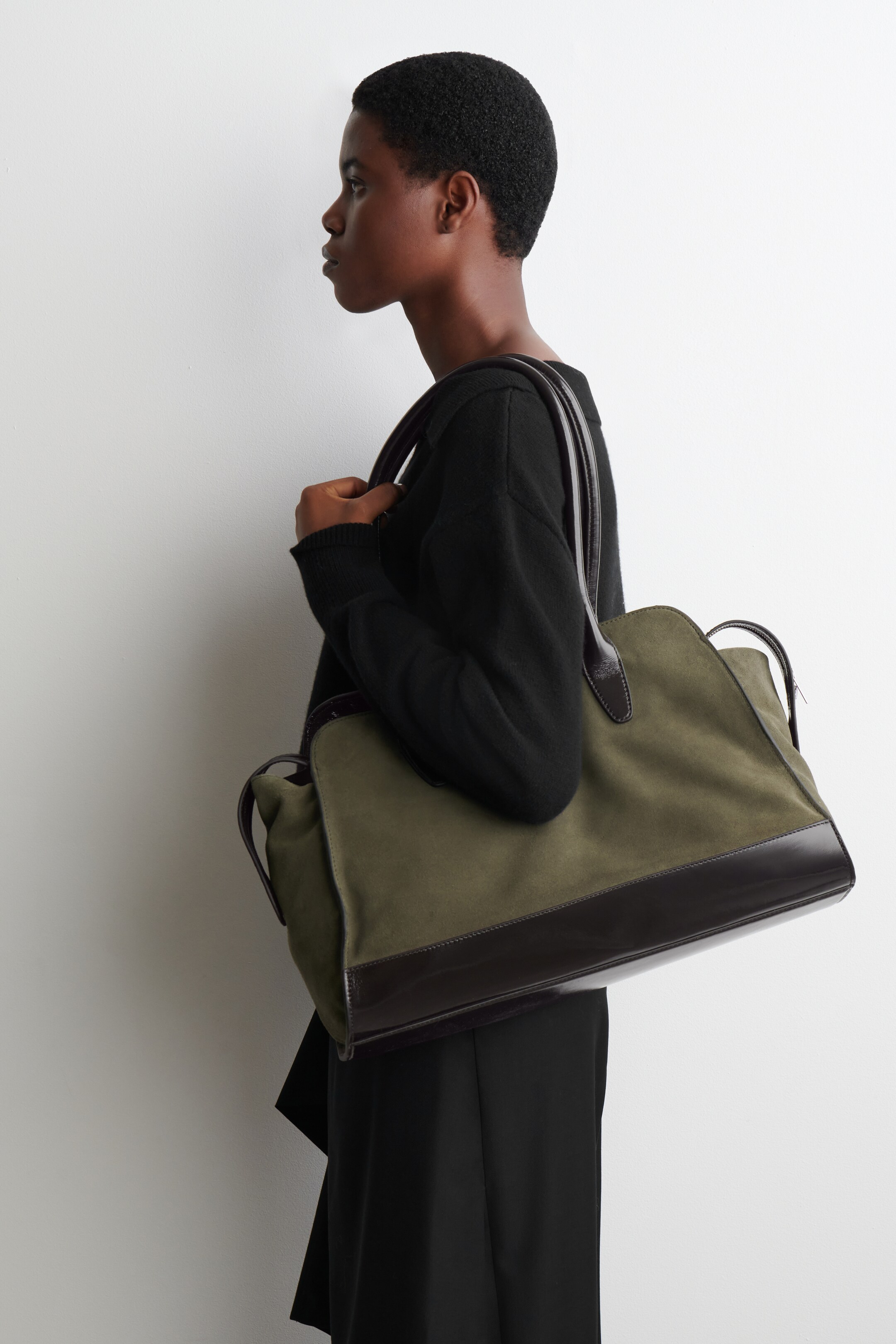 View larger image: A generous khaki suede tote bag, featuring a dark brown leather base and rounded handles, is being carried by the model. The model is holding the bag with their right hand, while its handles are resting on their right shoulder.