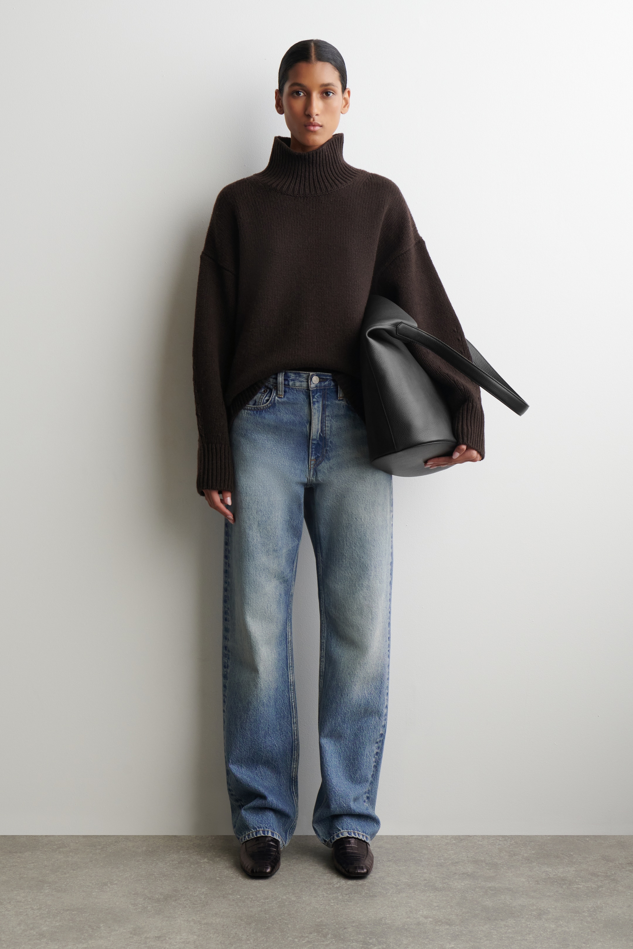 Agrandir l'image: Un pull à col roulé oversize marron foncé, avec un col roulé côtelé et de longues poignets côtelés, est porté avec un jean bleu à taille haute, à jambes larges et délavé clair. Un sac fourre-tout noir non structuré est tenu dans la main gauche, et des chaussures marron foncé à enfiler complètent l'ensemble.