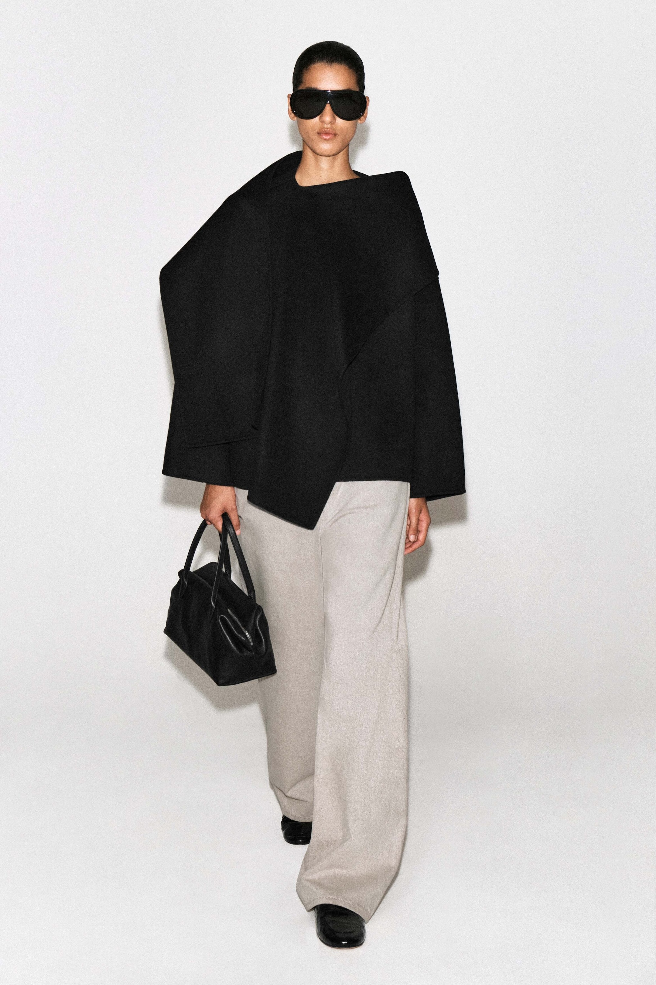 The double-faced wool-blend scarf jacket is shown from the front, featuring a relaxed, boxy silhouette in black with an integrated scarf draped over the shoulder. The jacket is designed with wide sleeves and an asymmetrical front panel that falls to hip length over light gray wide-leg trousers.