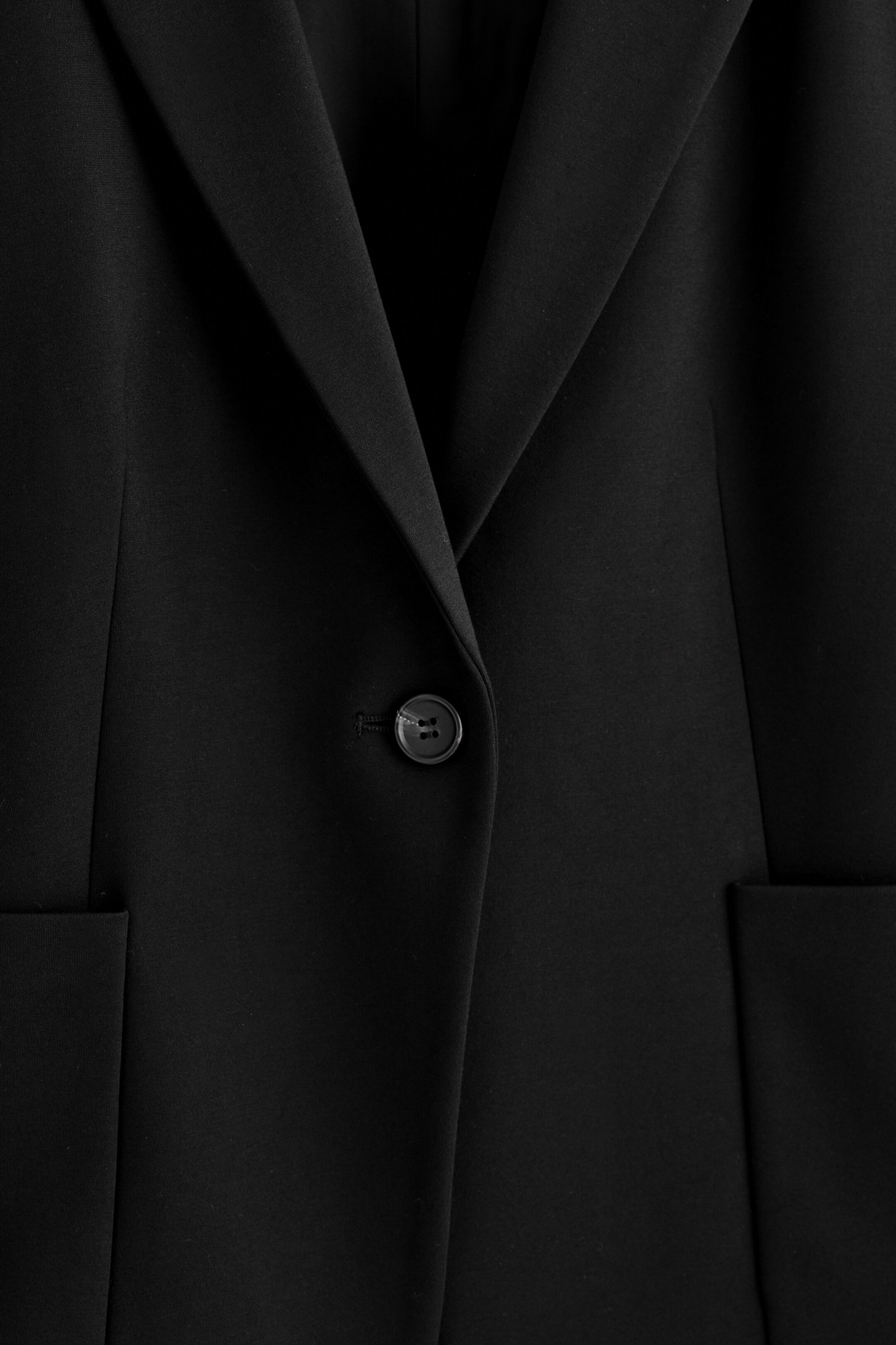 Zobrazit větší obrázek: Černé sako zobrazuje svou přední část, která obsahuje jedno černé čtyřdírkové zapínání na knoflík, klopový límec a dvě částečně viditelné kapsy s klopou.