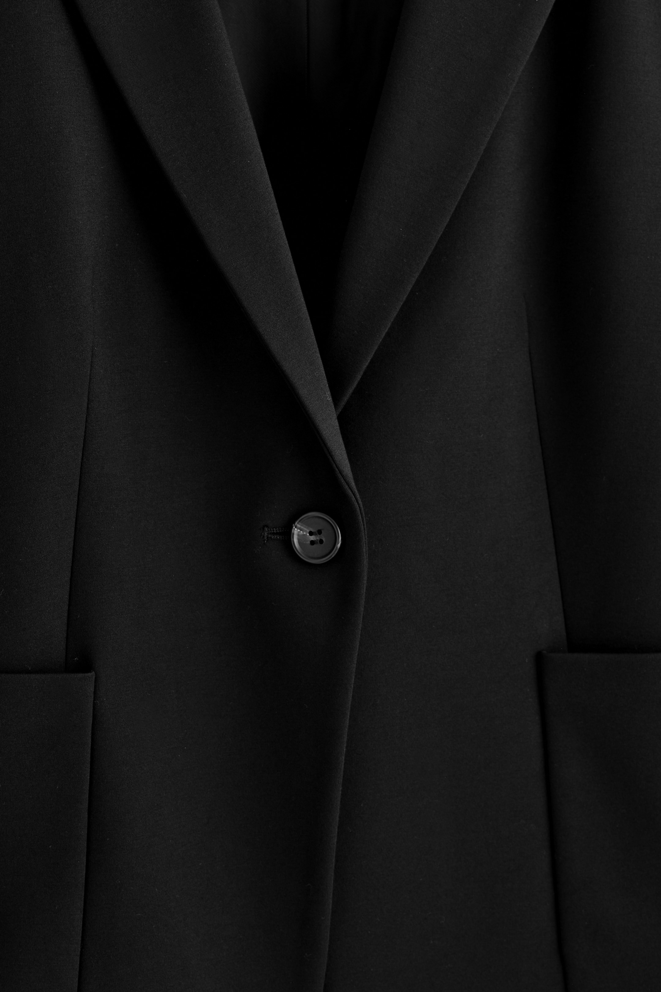 Agrandir l'image: Un blazer noir présente son devant, avec une fermeture à un bouton noir à quatre trous, un revers cranté et deux poches à rabat partiellement visibles.