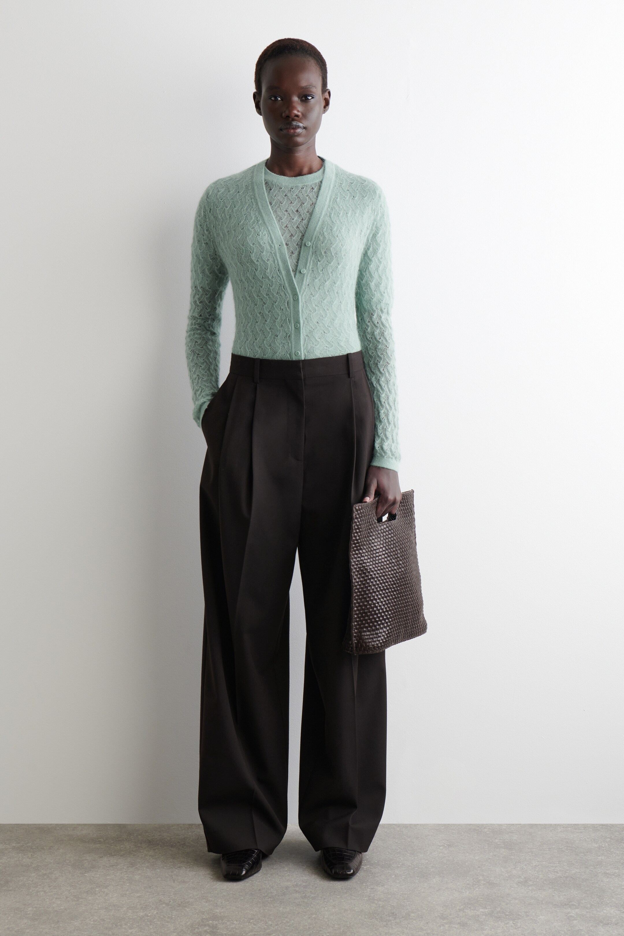 Größeres Bild anzeigen: Ein nach vorne gerichtetes Outfit wird präsentiert, das ein hellgrünes, strukturiertes Strick-Twinset mit einer V-Ausschnitt-Cardigan über einem passenden Rundhalstop zeigt. Dazu werden dunkelbraune, hoch taillierte, weit geschnittene Faltenhosen und dunkle, eckige Schuhe kombiniert, wobei eine dunkelbraune, geflochtene Tragetasche gehalten wird.