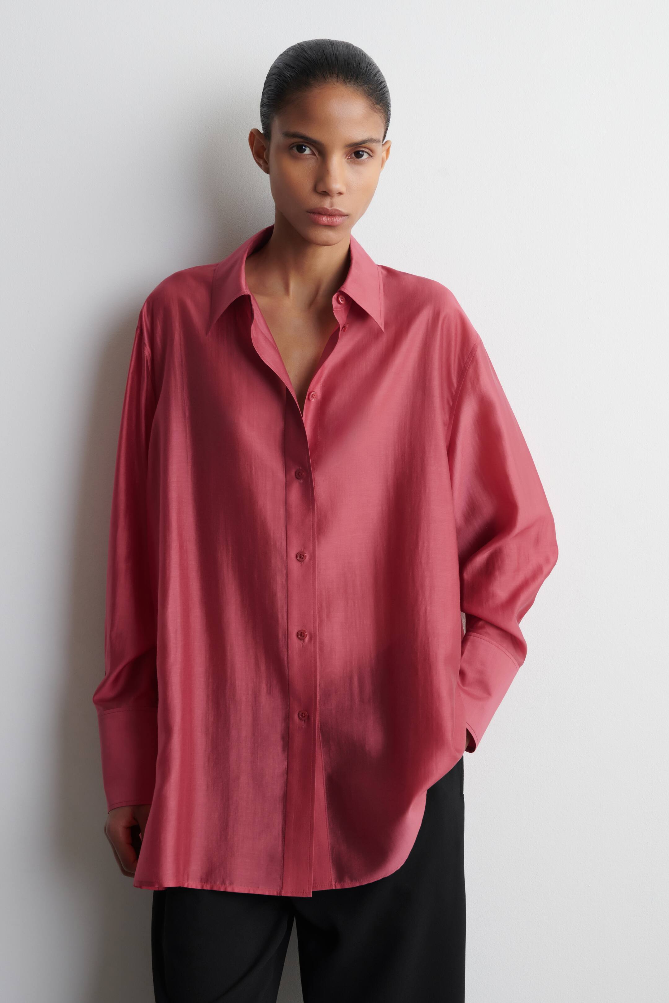 Agrandir l'image: Une chemise rose légèrement chatoyante et oversize est présentée de face, avec un col pointu et une patte de boutonnage complète avec des boutons assortis. Elle présente des manches longues avec des poignets larges et un ourlet arrondi, portée sur un pantalon noir.