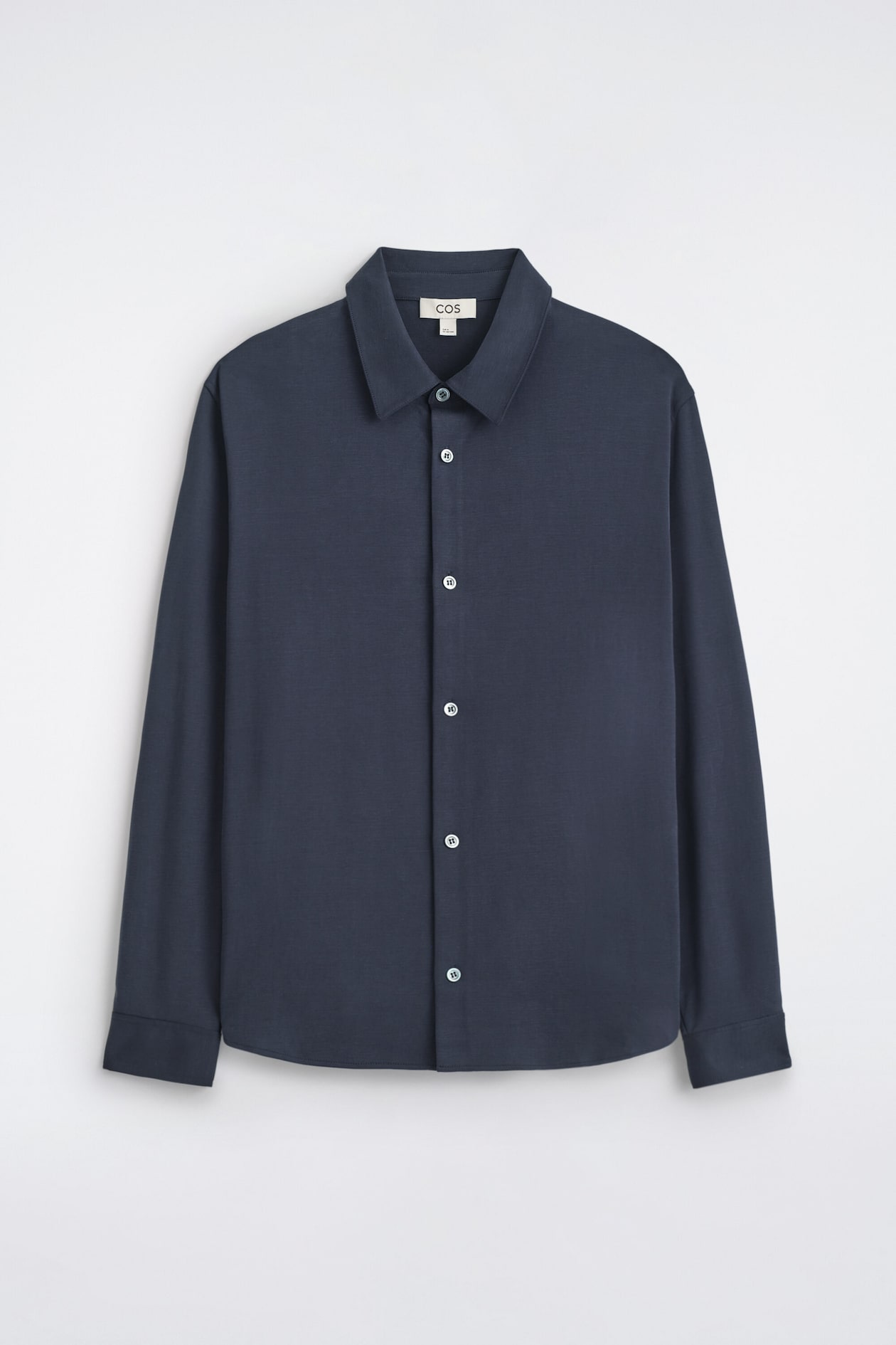 The FLUID INTERLOCK JERSEY SHIRT is shown in a front view, featuring a dark navy blue color, a classic pointed collar, and a central button-down closure with contrasting light buttons. It displays long sleeves with simple cuffs and a straight, clean-cut hem.