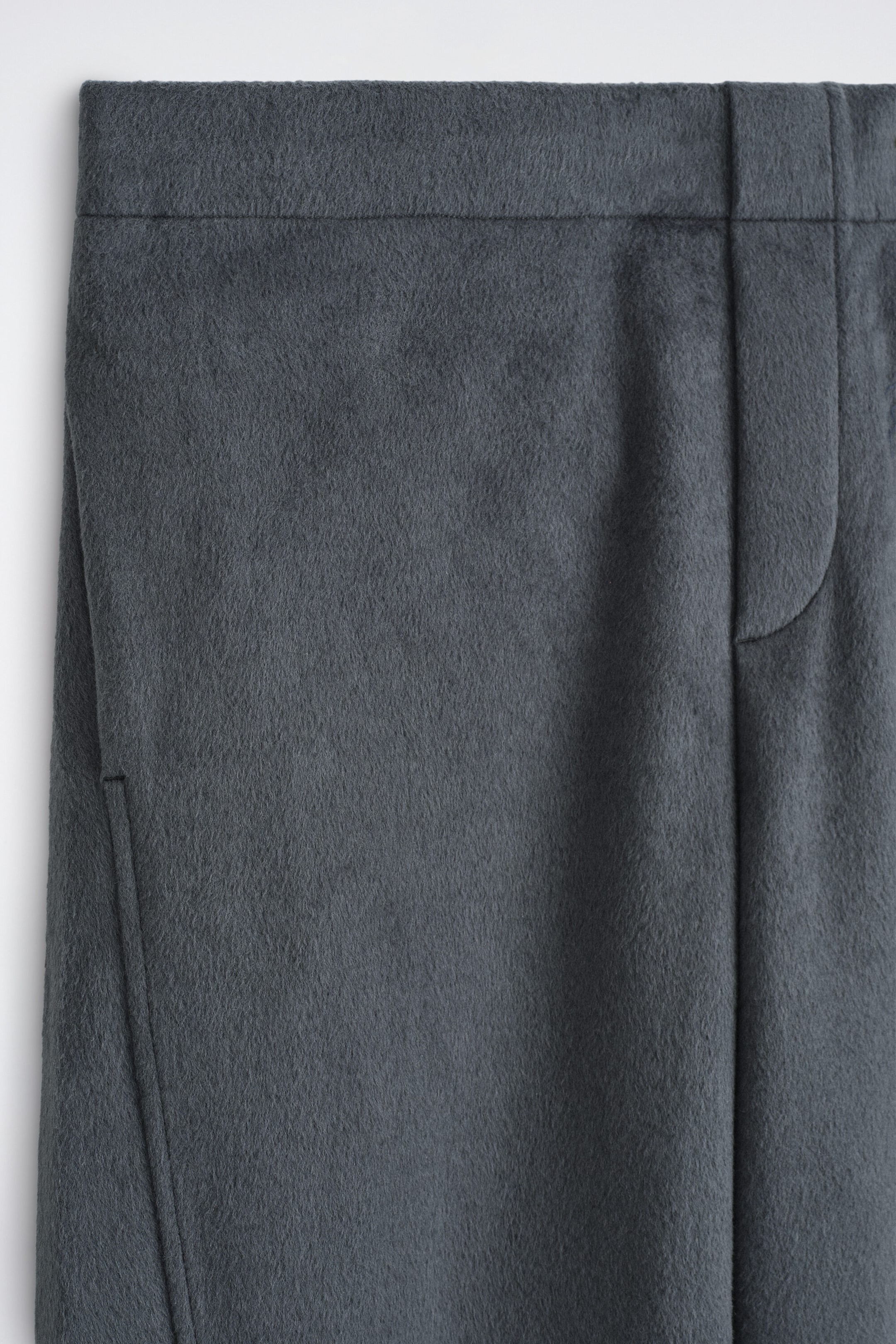 Un pantalon en laine brossée gris foncé est présenté en gros plan de face, avec une ceinture plate, une braguette dissimulée et une couture discrète de poche sur le côté.