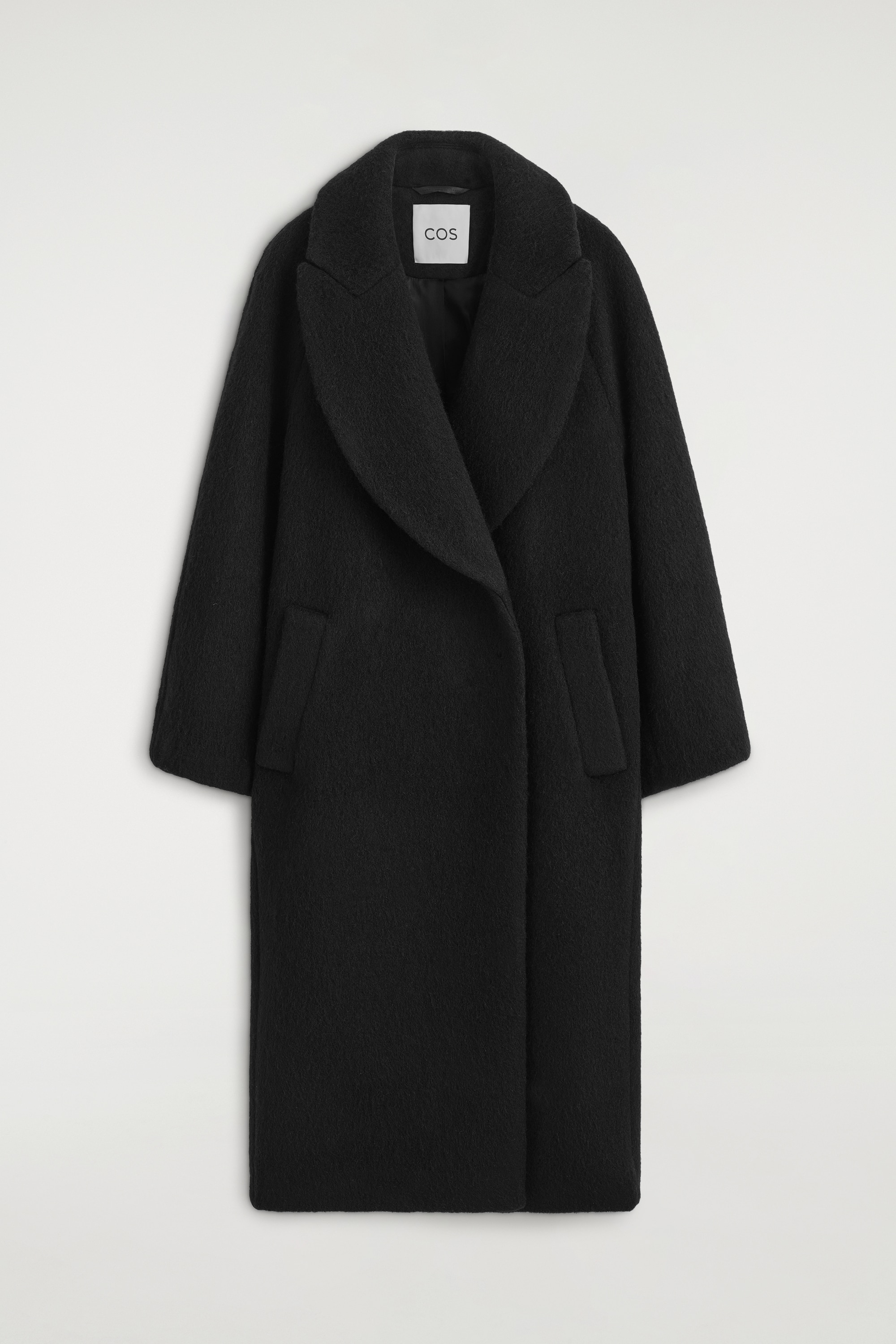 A black textured wool maxi coat featuring oversized lapels and a relaxed silhouette is being worn by the model. The coat is styled with a large black leather handbag, sheer black tights, and black pointed-toe shoes.