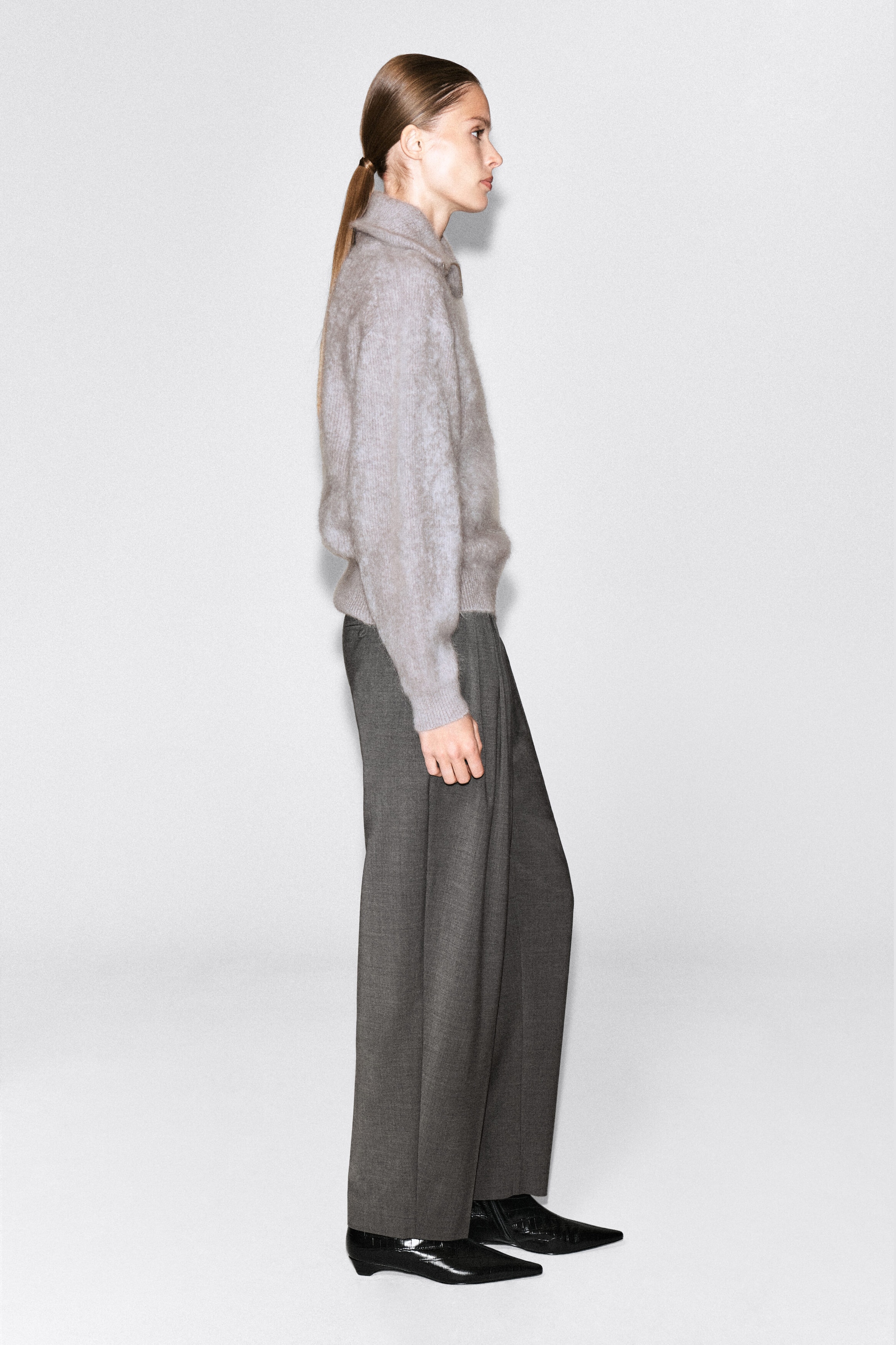 The model is wearing a light gray brushed-knit jumper featuring a high collar, long sleeves, and ribbed cuffs. Shown in side profile, the model styles the jumper with dark gray wide-leg trousers and black pointed-toe shoes. The model is wearing a light gray brushed-knit jumper featuring a high collar, long sleeves, and ribbed cuffs. Shown in side profile, the model styles the jumper with dark gray wide-leg trousers and black pointed-toe shoes.