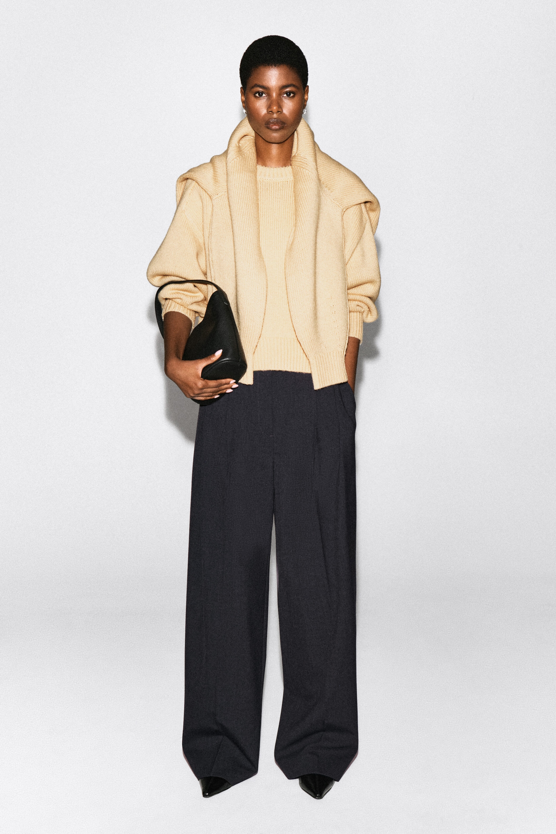 Un chandail à col rond en laine beige et aux lignes arrondies est porté par le mannequin, agencé avec un cardigan en tricot assorti drapé sur les épaules et un pantalon large foncé. Le mannequin tient un sac à main en cuir noir.