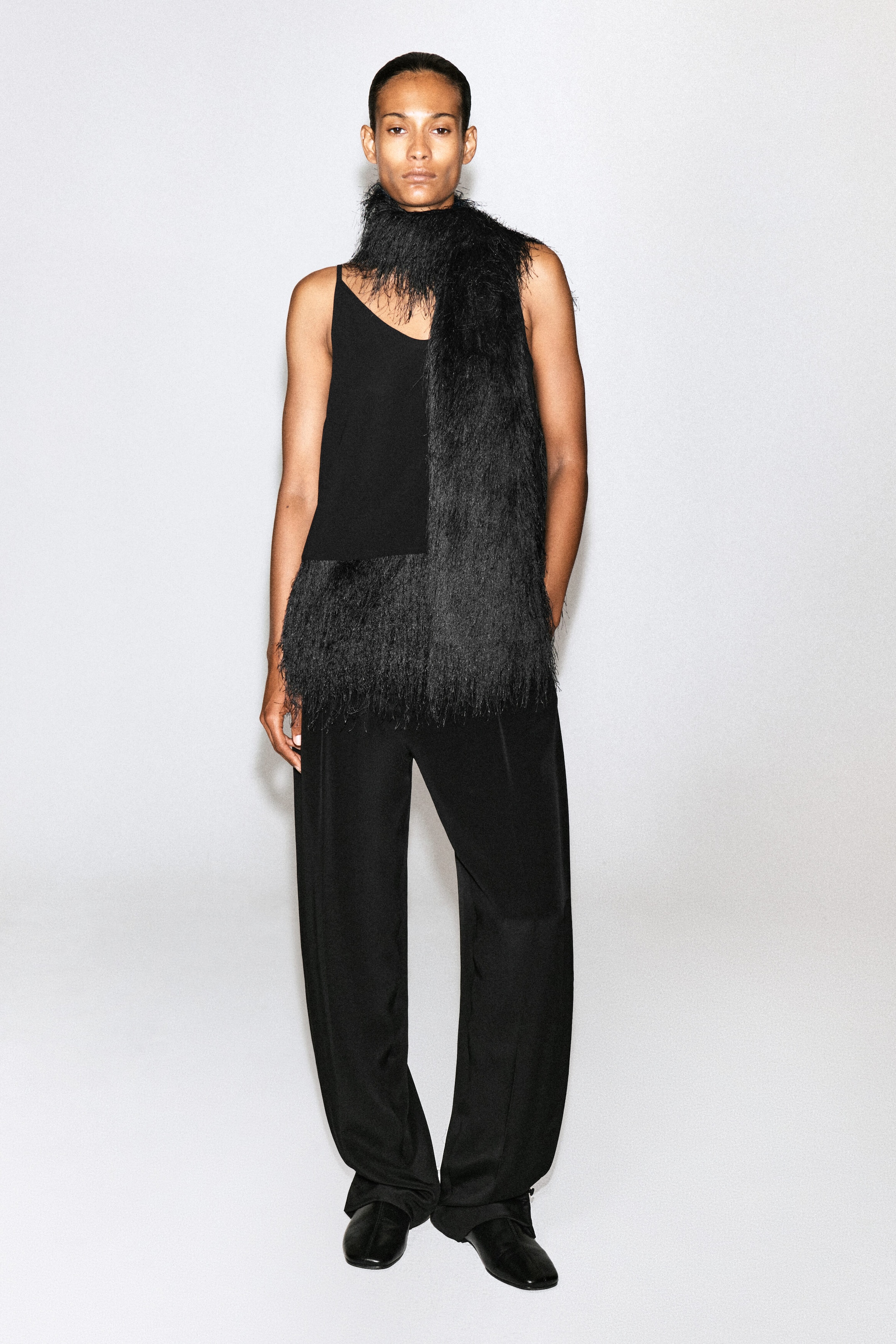 A black scarf featuring long, wispy fringe is being worn by the model, wrapped around the neck with one end hanging down the front of the body. The scarf is styled over a black sleeveless top and trousers.