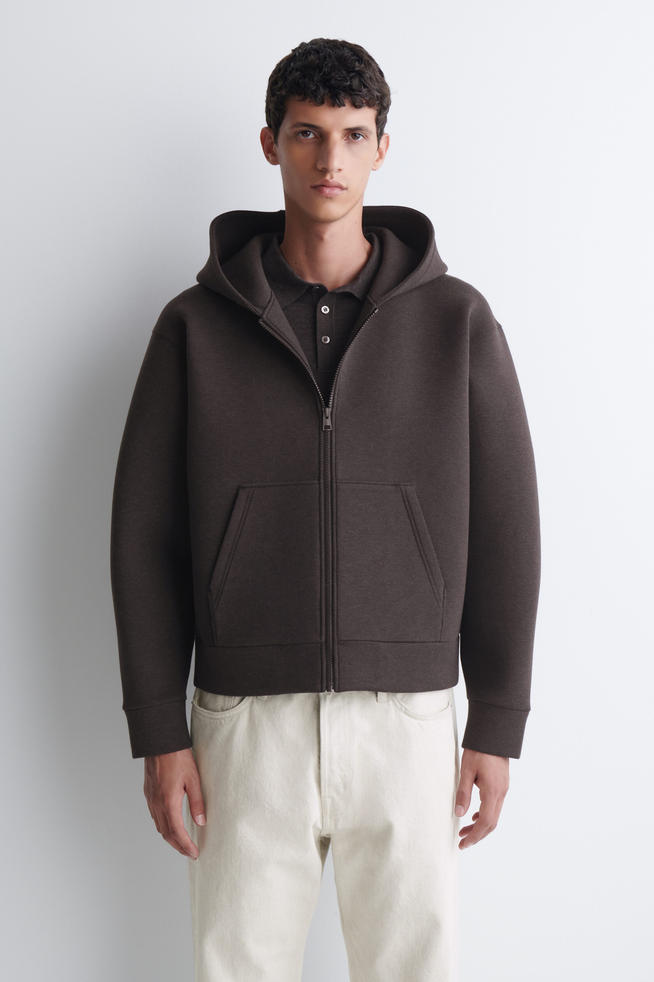An oversized scuba jersey zip-up hoodie in dark brown is worn by the model, featuring a structured hood, a front zipper, and a split kangaroo pocket. The hoodie is layered over a matching dark brown polo shirt and paired with off-white trousers. An oversized scuba jersey zip-up hoodie in dark brown is worn by the model, featuring a structured hood, a front zipper, and a split kangaroo pocket. The hoodie is layered over a matching dark brown polo shirt and paired with off-white trousers.