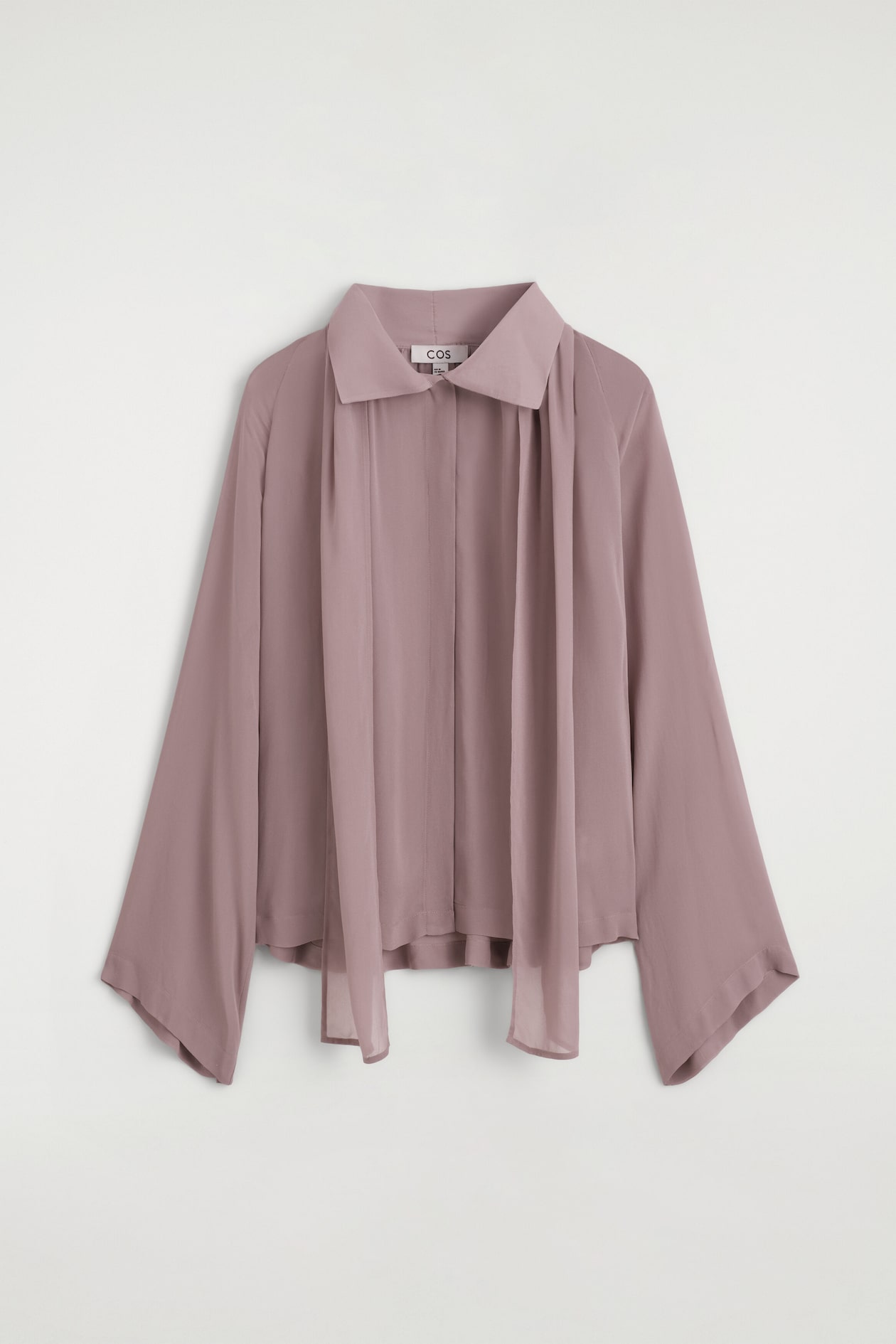 Le haut fluide à détail foulard est présenté de face dans une nuance mauve poudré, avec un col pointu et deux longs pans assortis en tissu tombant sur le devant de la poitrine. La blouse présente une silhouette décontractée avec des manches larges et fluides ainsi qu’une patte de boutonnage dissimulée sur le devant.
