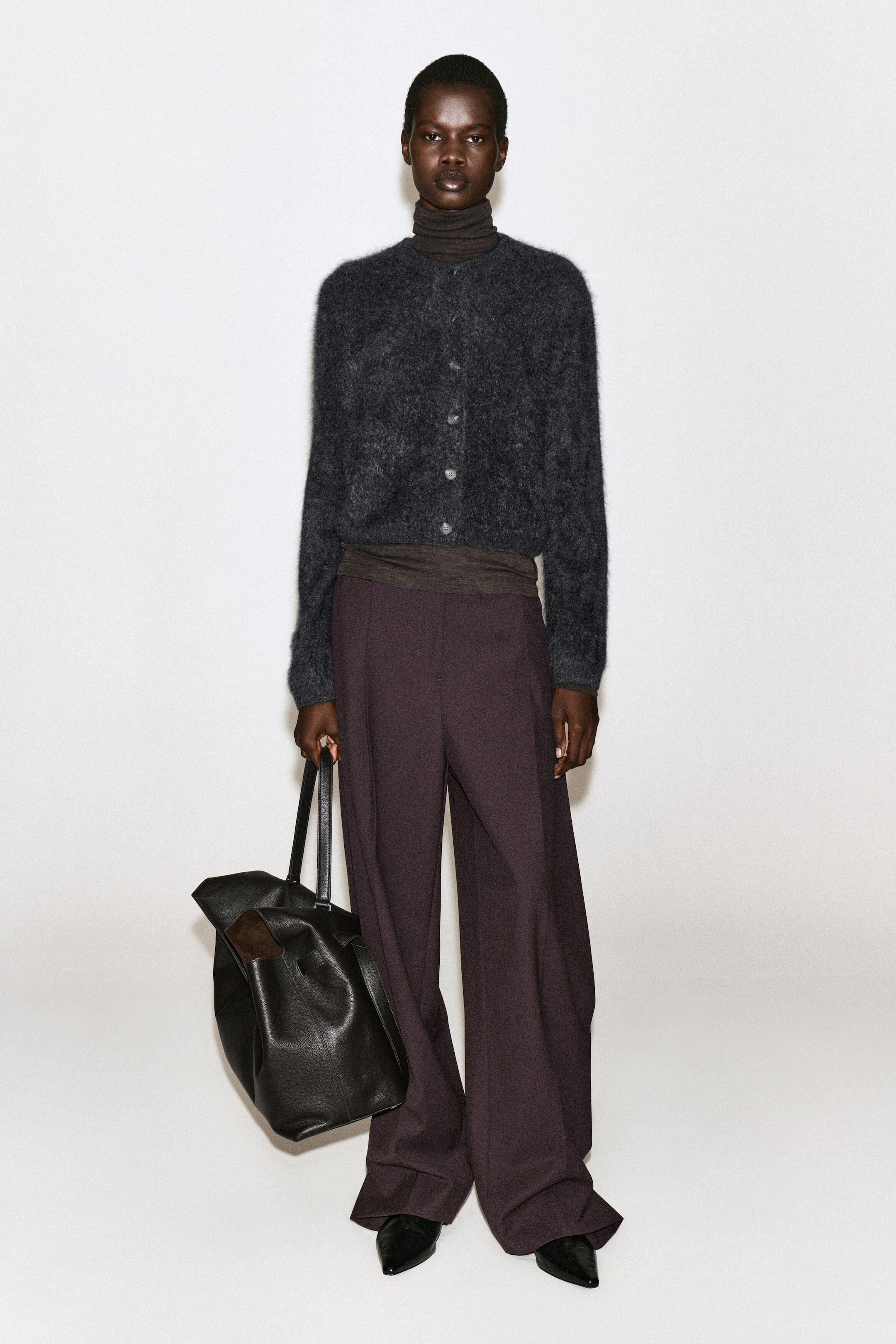 A dark grey, brushed cashmere crew-neck cardigan is worn buttoned up by the model over a dark brown turtleneck. The fuzzy-textured cardigan is styled with dark purple wide-leg trousers, and the model is holding a large black leather tote bag. A dark grey, brushed cashmere crew-neck cardigan is worn buttoned up by the model over a dark brown turtleneck. The fuzzy-textured cardigan is styled with dark purple wide-leg trousers, and the model is holding a large black leather tote bag.