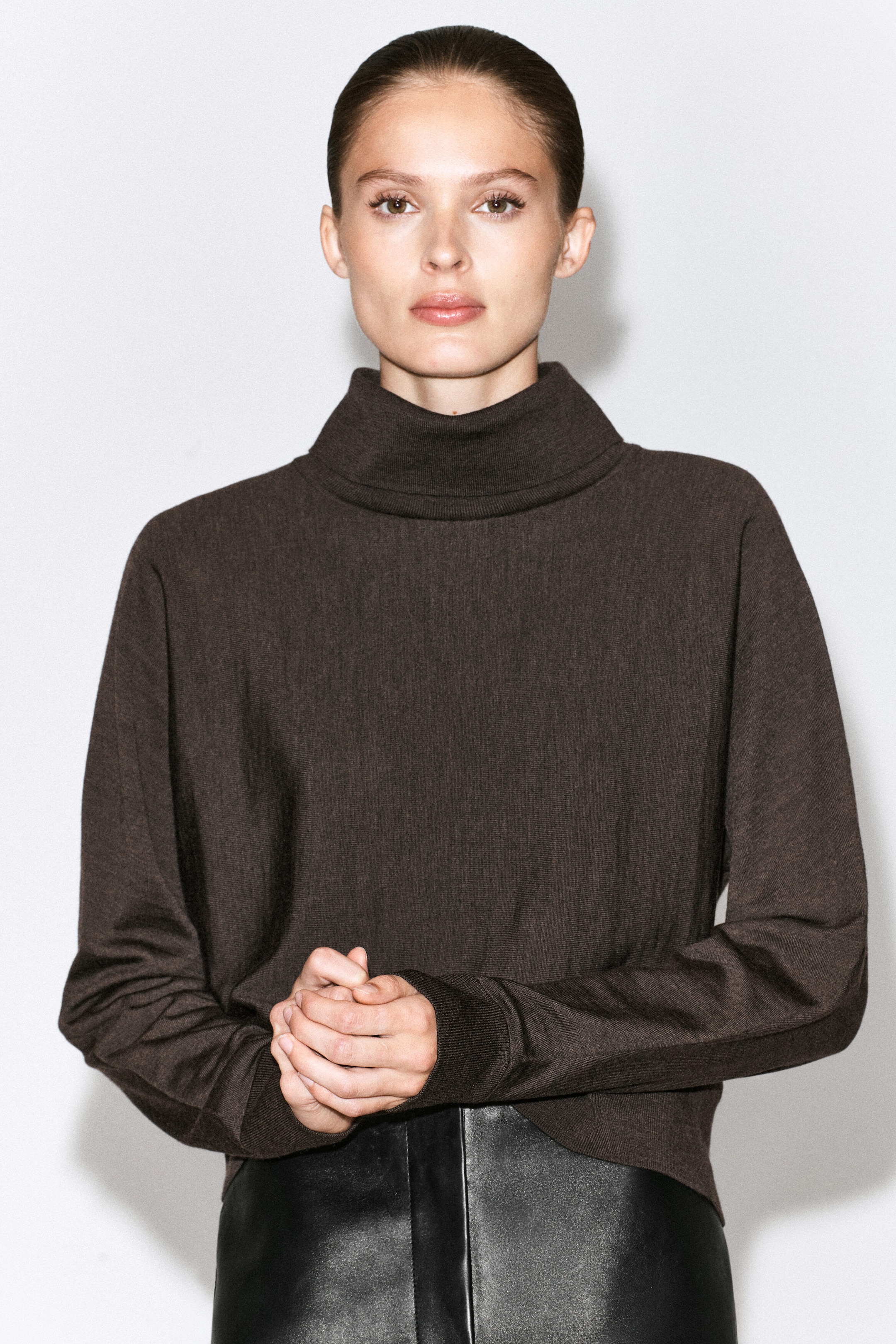 The model is wearing a dark brown long-sleeved roll-neck sweater, with their hands clasped together at the waist. The fine-knit sweater features a high rolled collar and ribbed cuffs, and is styled with black leather trousers. The model is wearing a dark brown long-sleeved roll-neck sweater, with their hands clasped together at the waist. The fine-knit sweater features a high rolled collar and ribbed cuffs, and is styled with black leather trousers.