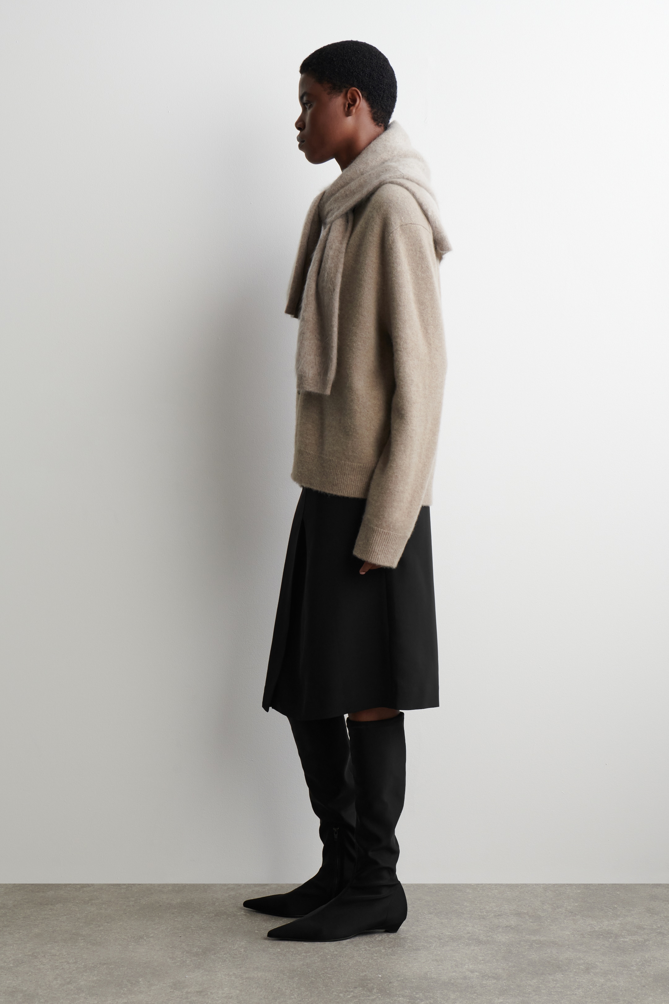 Grösseres Bild ansehen: Ein hellbrauner, locker sitzender Pullover mit gerippten Bündchen wird im Profil präsentiert, mit einem passenden Schal, der locker über die Schultern drapiert ist. Darunter wird ein schwarzer Midirock mit einer dezenten Frontfalte gezeigt. Schwarze, spitz zulaufende, flache Overknee-Stiefel vervollständigen das Ensemble.