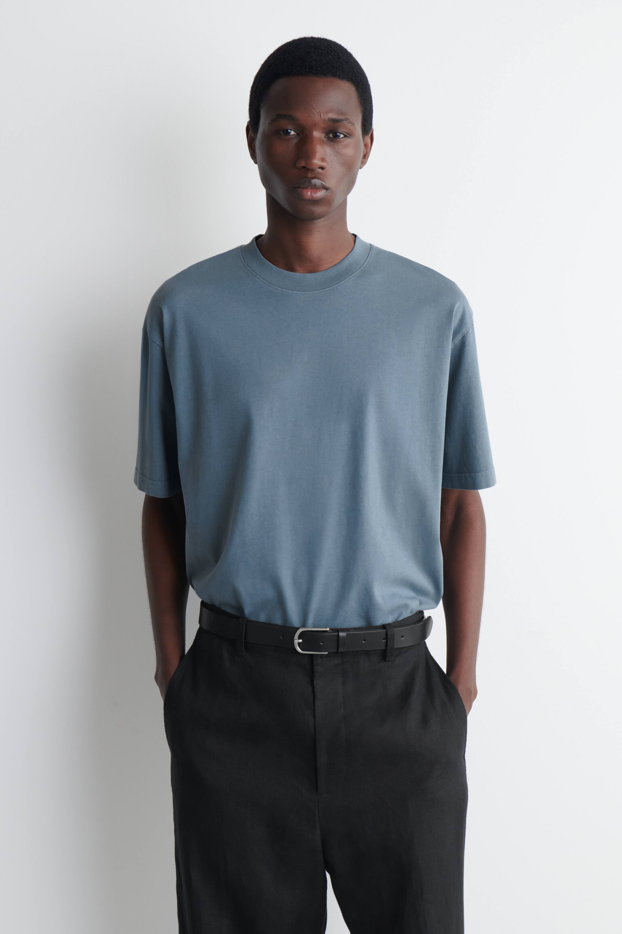 Se større billede: En skiferblå t-shirt bæres, med korte ærmer og en ribbet rund hals. Den præsenterer en oversized pasform, mens den er stukket ned i sorte bukser.