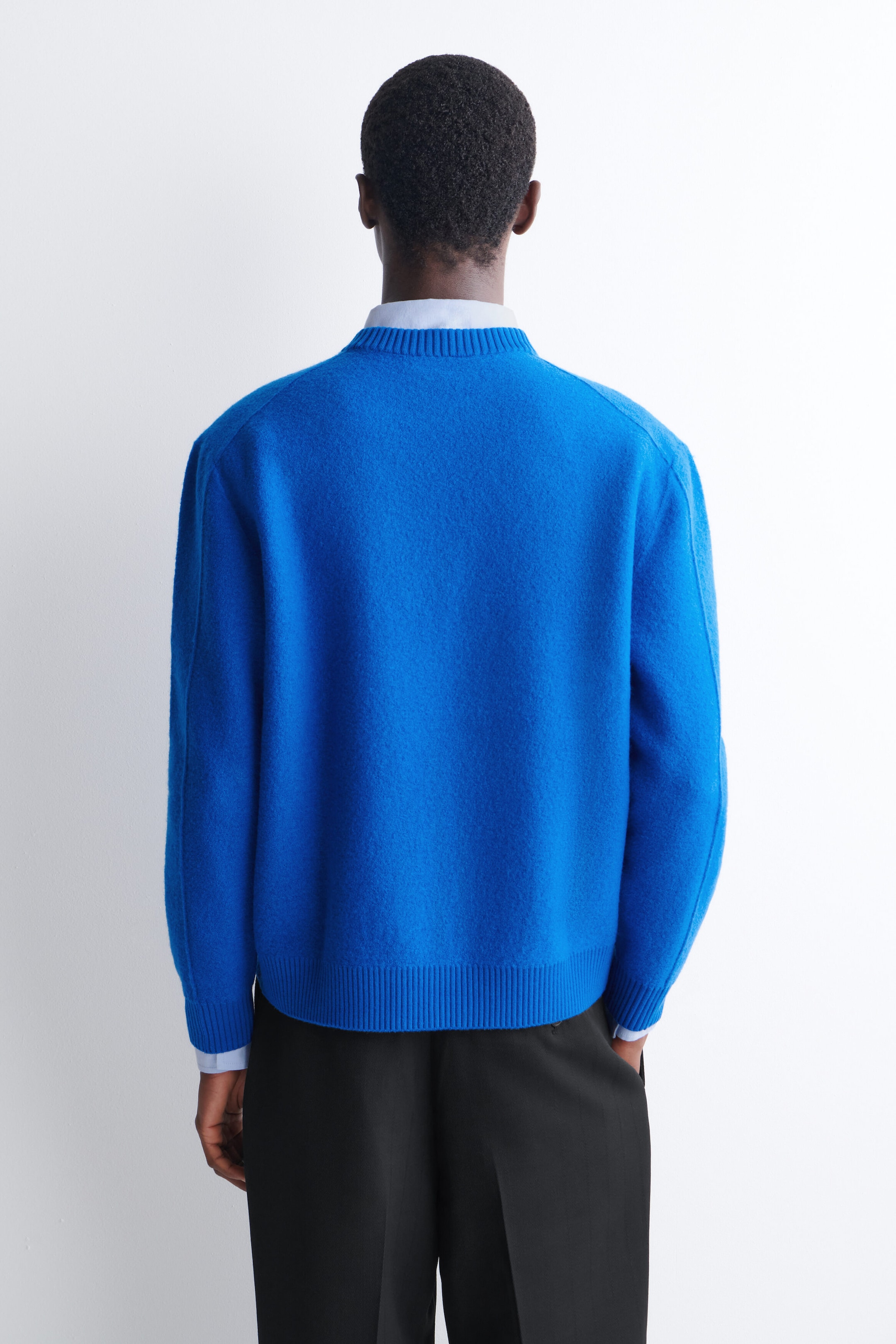 A bright blue boiled-wool sweater with a ribbed collar, hem, and cuffs is worn by the model, shown from the back. The sweater is layered over a light blue collared shirt and paired with black trousers. A bright blue boiled-wool sweater with a ribbed collar, hem, and cuffs is worn by the model, shown from the back. The sweater is layered over a light blue collared shirt and paired with black trousers.