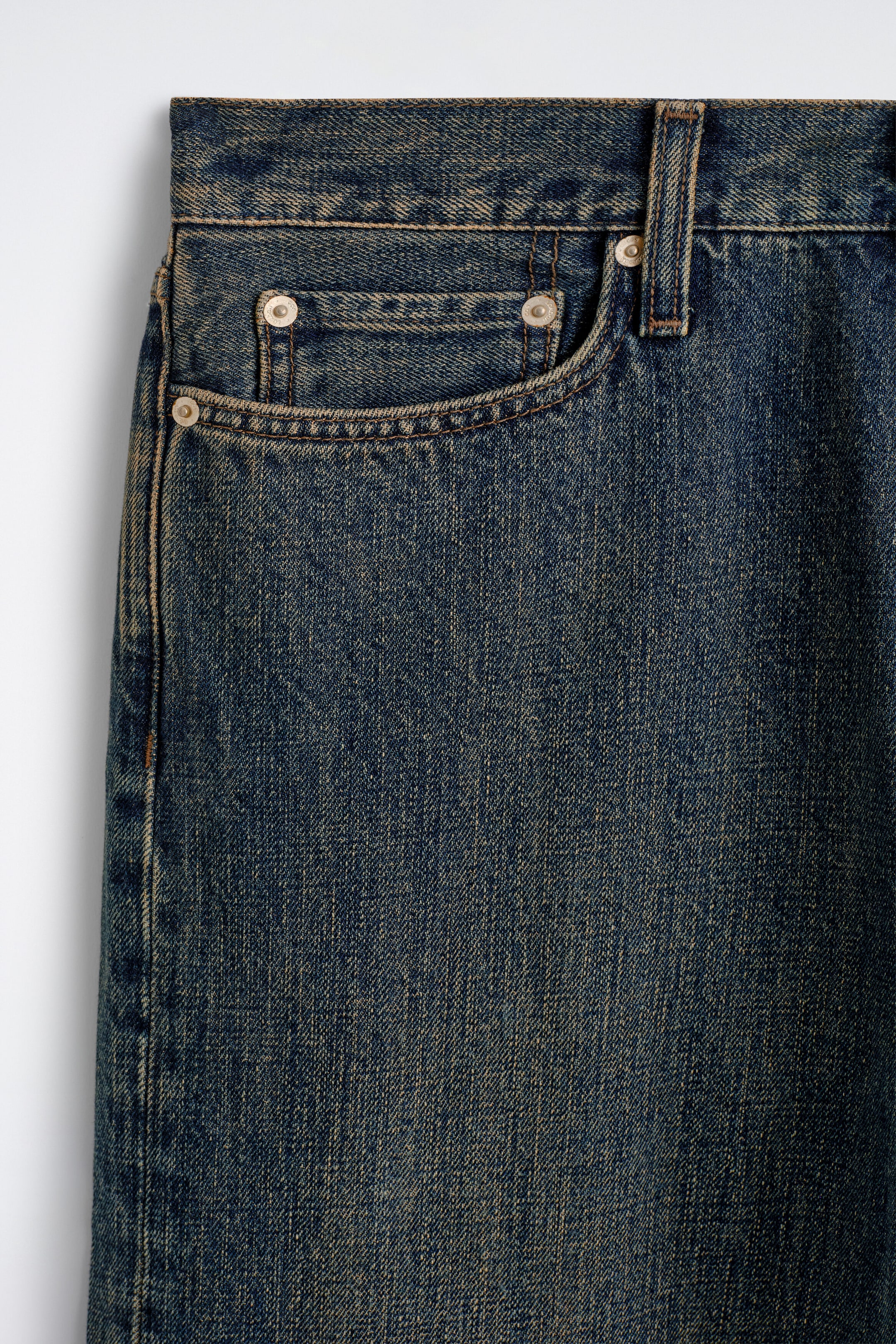 A close-up of the front right side of a pair of dark blue denim jeans showing the curved front pocket and a smaller coin pocket secured with metallic rivets. The denim features a textured wash with gold-toned contrast stitching along the seams, a belt loop, and the waistband. A close-up of the front right side of a pair of dark blue denim jeans showing the curved front pocket and a smaller coin pocket secured with metallic rivets. The denim features a textured wash with gold-toned contrast stitching along the seams, a belt loop, and the waistband.