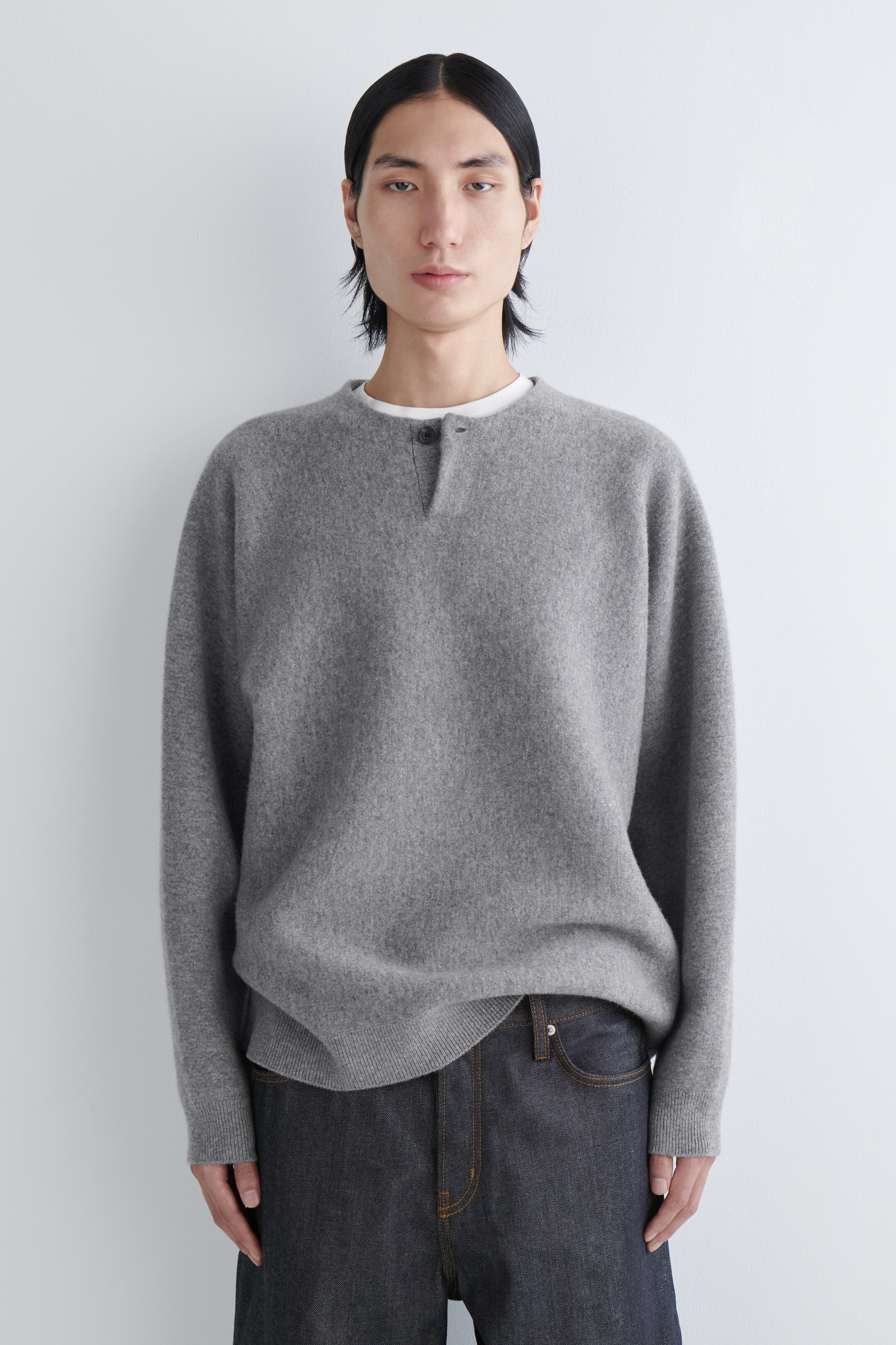 Agrandir l'image: Un pull henley gris est présenté de face, mettant en valeur une texture tricotée douce et duveteuse et une patte de boutonnage à un bouton au niveau de l'encolure. Il présente une coupe décontractée avec un ourlet côtelé, et un t-shirt à col rond blanc est visible en dessous.