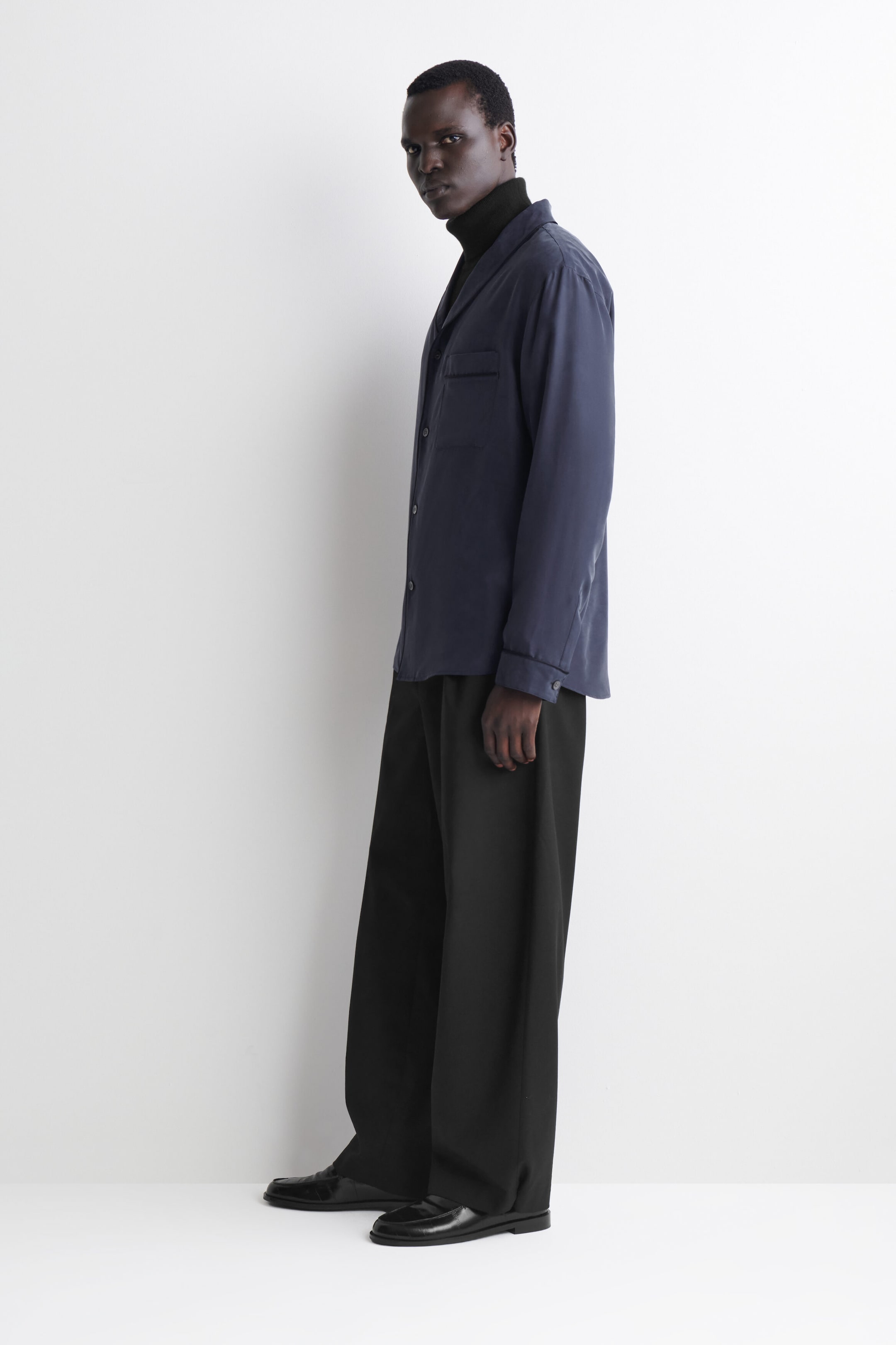 The navy blue silk shirt is shown on a model facing left, showcasing its relaxed, fluid drape. The shirt features a shawl collar, subtle piping details on the chest pocket and cuffs, and is layered over a black roll-neck top. The navy blue silk shirt is shown on a model facing left, showcasing its relaxed, fluid drape. The shirt features a shawl collar, subtle piping details on the chest pocket and cuffs, and is layered over a black roll-neck top.