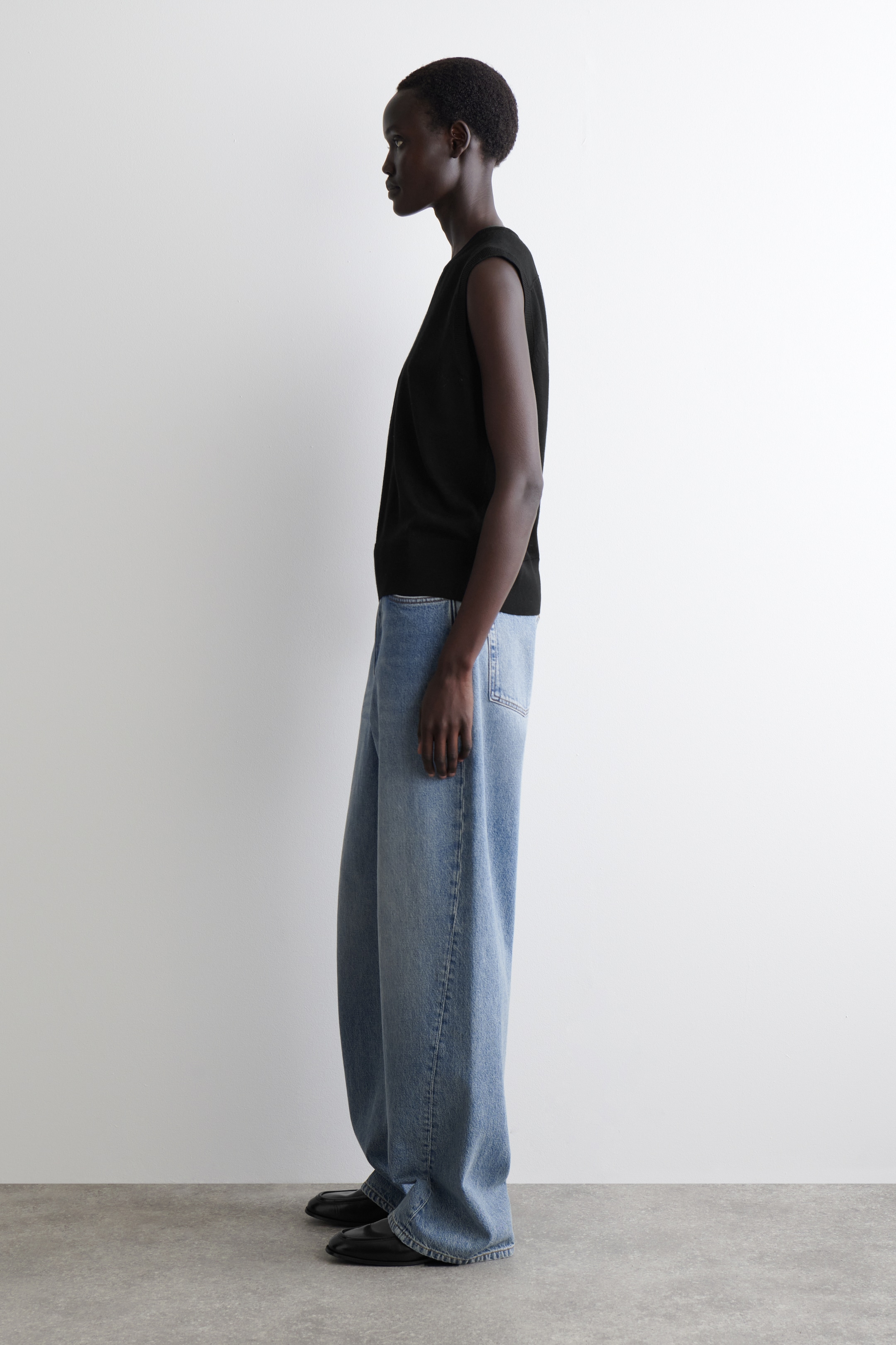 Visualizza immagine più grande: Vista di lato, una canotta nera senza maniche è indossata con jeans in denim azzurro chiaro a gamba larga e scarpe basse nere. La canotta presenta una vestibilità rilassata, e i jeans mostrano una silhouette ampia e a tutta lunghezza che si adagia leggermente sulle calzature.