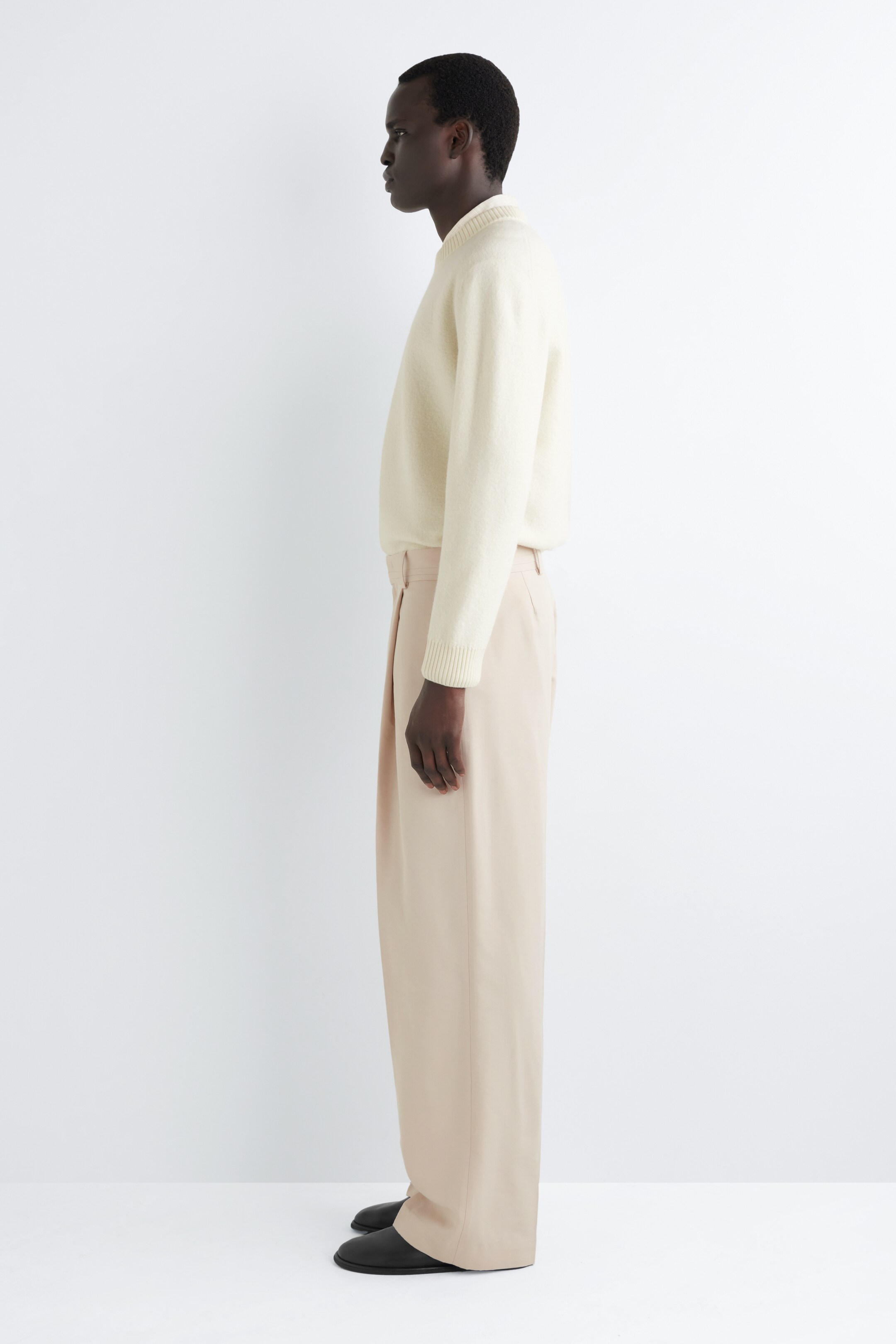 Agrandir l'image: Pantalon large de couleur beige clair avec une coupe décontractée, des plis profonds à la taille et des passants de ceinture visibles. Le pantalon, incliné vers la gauche, présente un pli central net descendant chaque jambe et un drapé fluide.