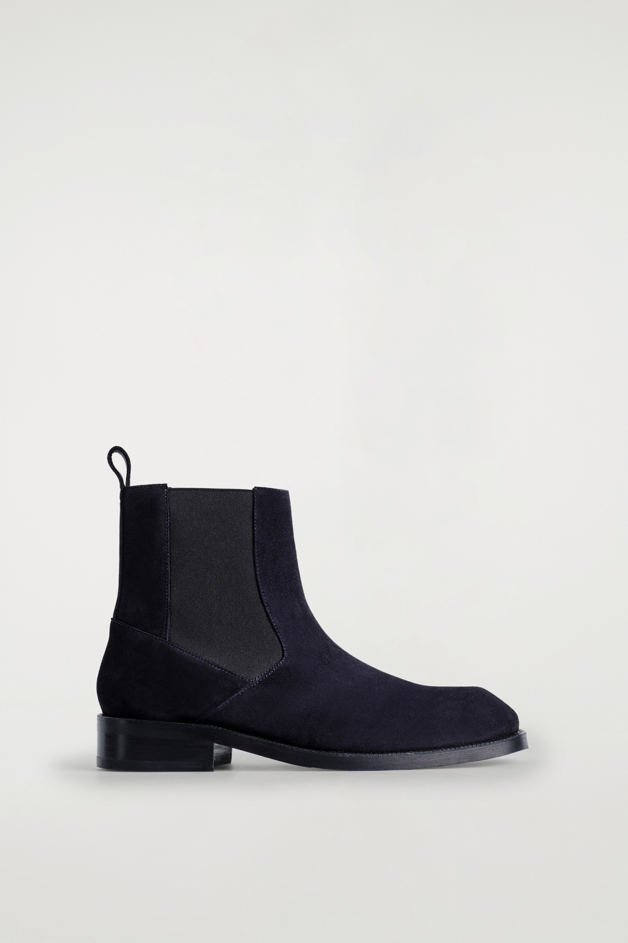 Grotere afbeelding bekijken: Een donker suède leren Chelsea-boot wordt getoond met de voorkant naar rechts, met een eigentijdse vierkante neus, een elastische zij-inzet en een treklus aan de achterkant van de enkel.