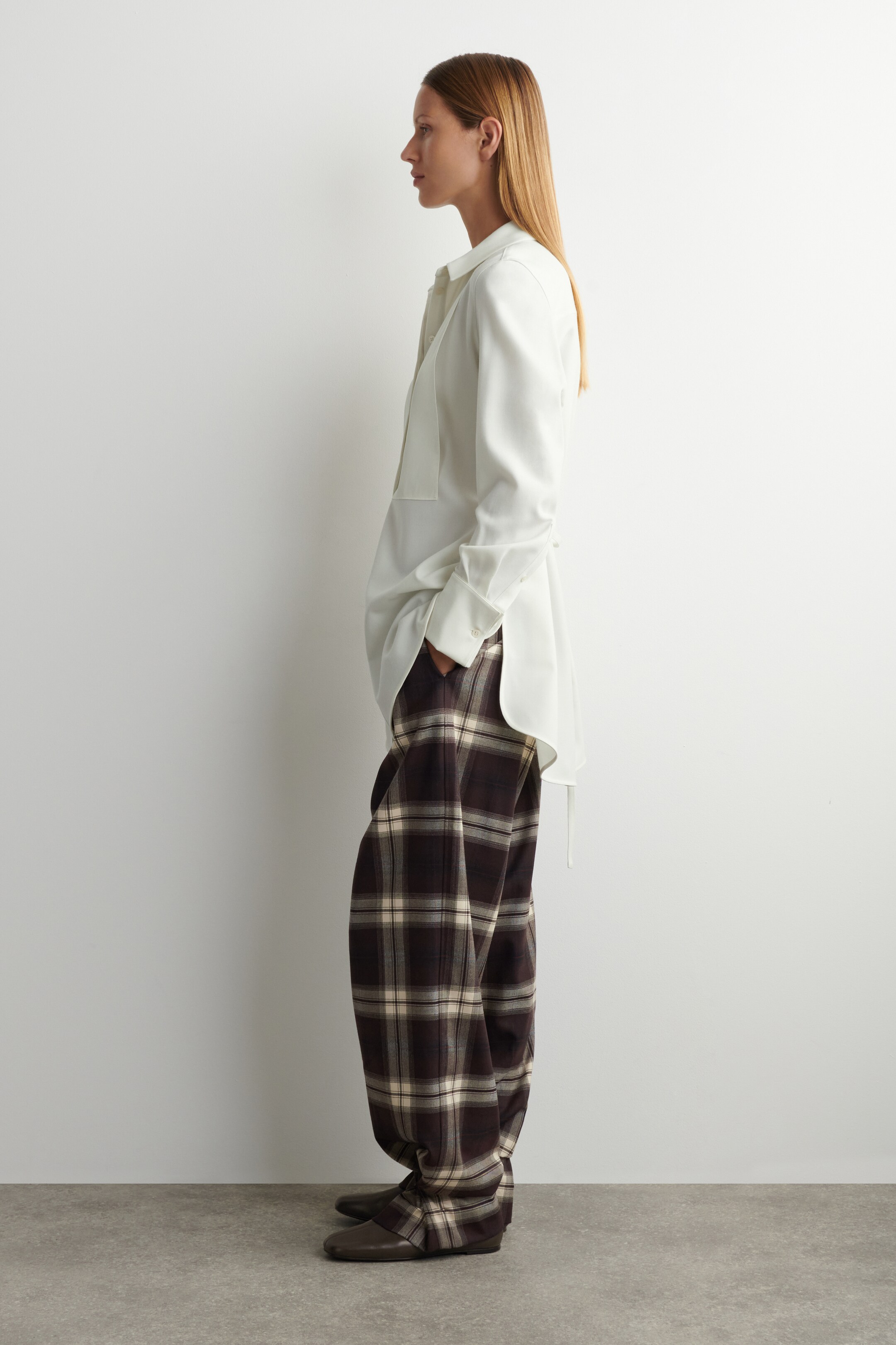 Agrandir l'image: Le modèle porte un pantalon large à carreaux, avec un motif à carreaux brun foncé et crème. Une chemise de couleur claire, à coupe décontractée, est superposée sur le pantalon, avec la main du modèle placée dans une poche du pantalon, et des chaussures plates et sombres aux pieds.