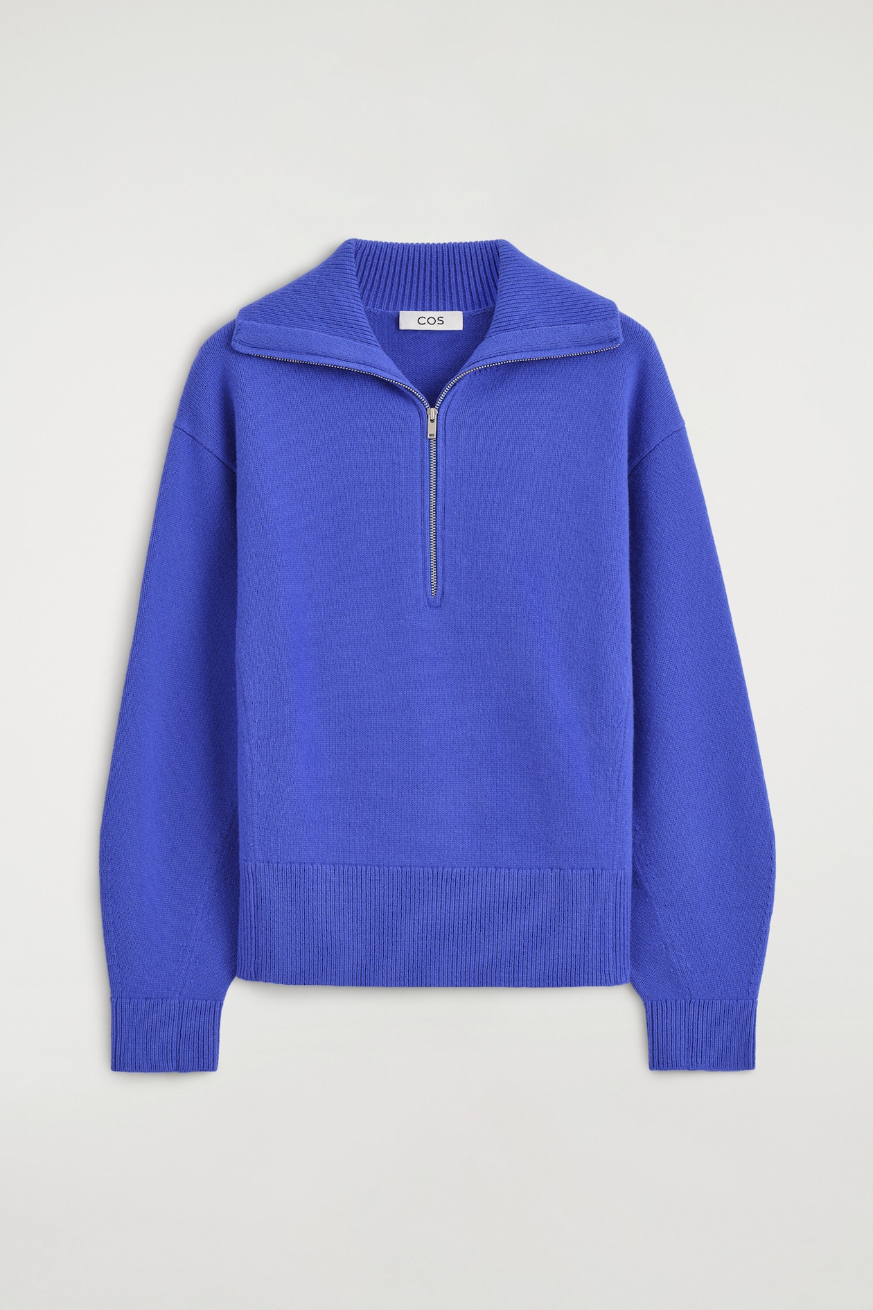 A blue knit sweater is shown from the front, featuring a relaxed fit with dropped shoulders and long, sculptural sleeves. The garment features a silver-tone half-zip closure that opens to a wide, ribbed collar, and is finished with a ribbed hem and cuffs.