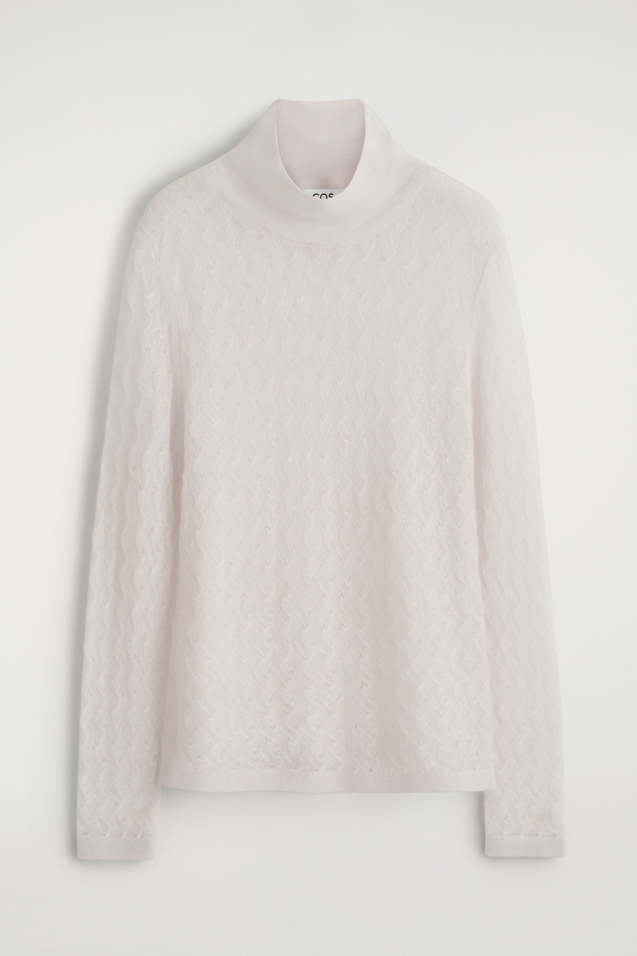 Visa större bild: En ljus off-white långärmad topp ligger platt och visar sin framsida, med en ribbad trattkrage och ett fint pointelle-stickmönster överallt som skapar en subtilt genomskinlig finish. Manschetterna och fållen är också ribbade.