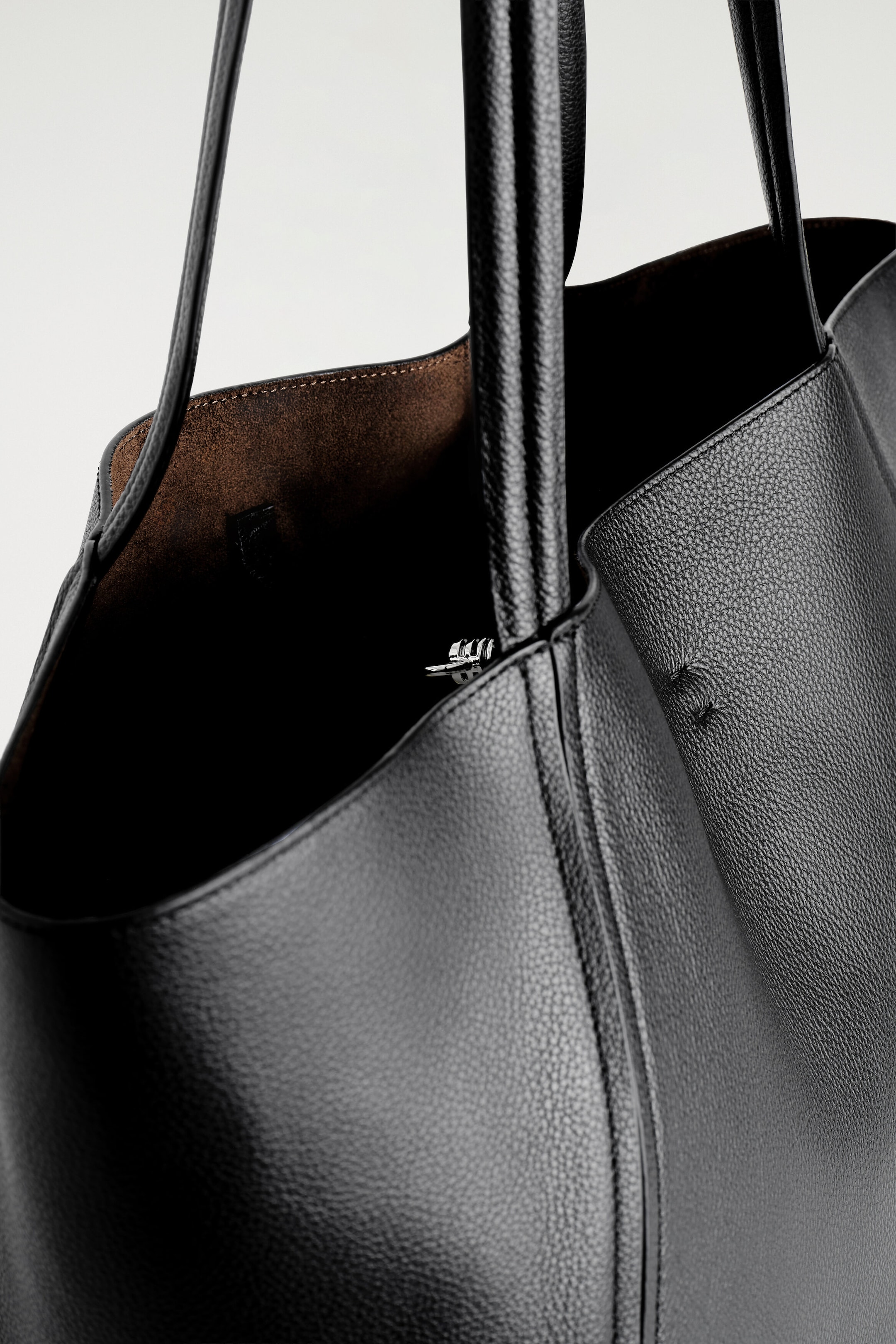 A black grained leather shopper bag is shown in a close-up, angled view, highlighting its textured surface and curved stitched seams. The open top reveals a brown bonded lining and a silver-tone metal clip closure. A black grained leather shopper bag is shown in a close-up, angled view, highlighting its textured surface and curved stitched seams. The open top reveals a brown bonded lining and a silver-tone metal clip closure.