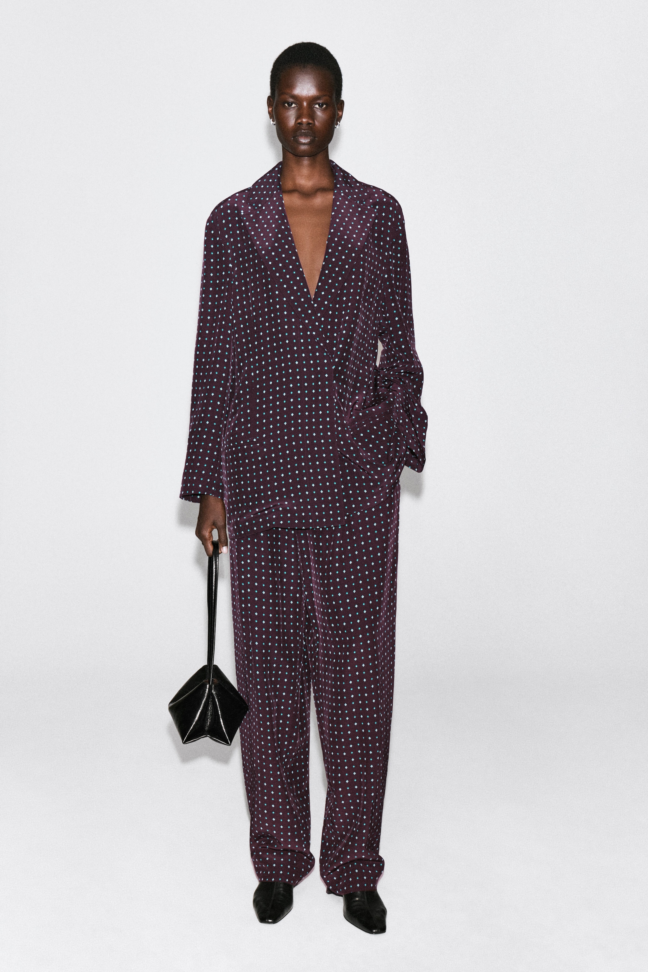 The diamond-print double-breasted blazer is shown from the front, featuring a relaxed fit in a deep purple fabric with a repeating pattern of small teal diamonds. It drapes loosely over the body with a deep V-neckline and is styled with matching trousers to create a coordinated suit. The diamond-print double-breasted blazer is shown from the front, featuring a relaxed fit in a deep purple fabric with a repeating pattern of small teal diamonds. It drapes loosely over the body with a deep V-neckline and is styled with matching trousers to create a coordinated suit.