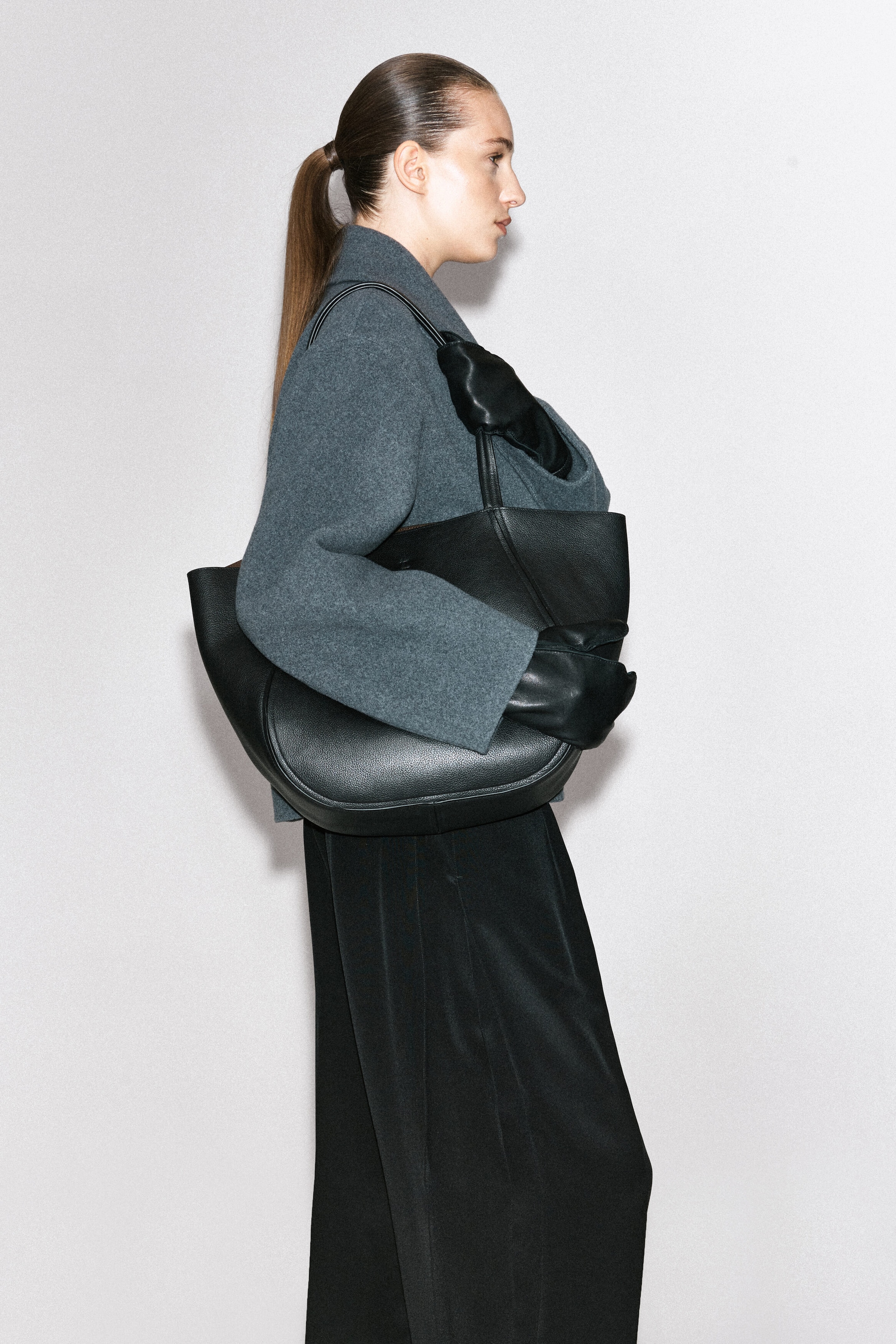 A black leather tote bag with a curved silhouette is carried under the model’s arm. Wearing black leather gloves and a grey wool coat, the model holds the bag’s shoulder strap and keeps the body of the bag tucked close at their side. A black leather tote bag with a curved silhouette is carried under the model’s arm. Wearing black leather gloves and a grey wool coat, the model holds the bag’s shoulder strap and keeps the body of the bag tucked close at their side.