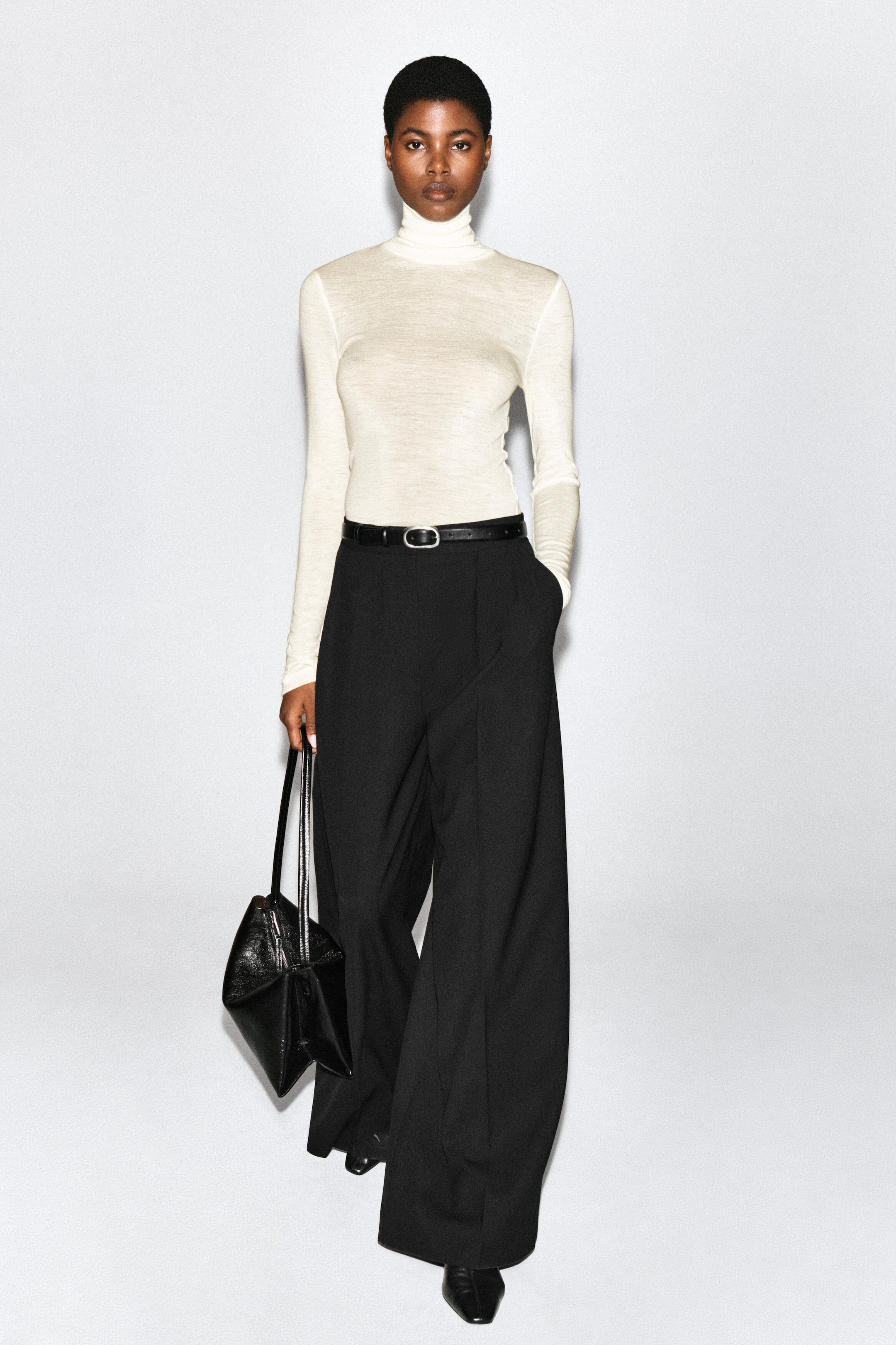 Pleated wide-leg pants in black are worn by the model, styled with a black leather belt and a tucked-in cream turtleneck top. The model is standing with one hand in the pants pocket while carrying a black leather handbag. Pleated wide-leg pants in black are worn by the model, styled with a black leather belt and a tucked-in cream turtleneck top. The model is standing with one hand in the pants pocket while carrying a black leather handbag.
