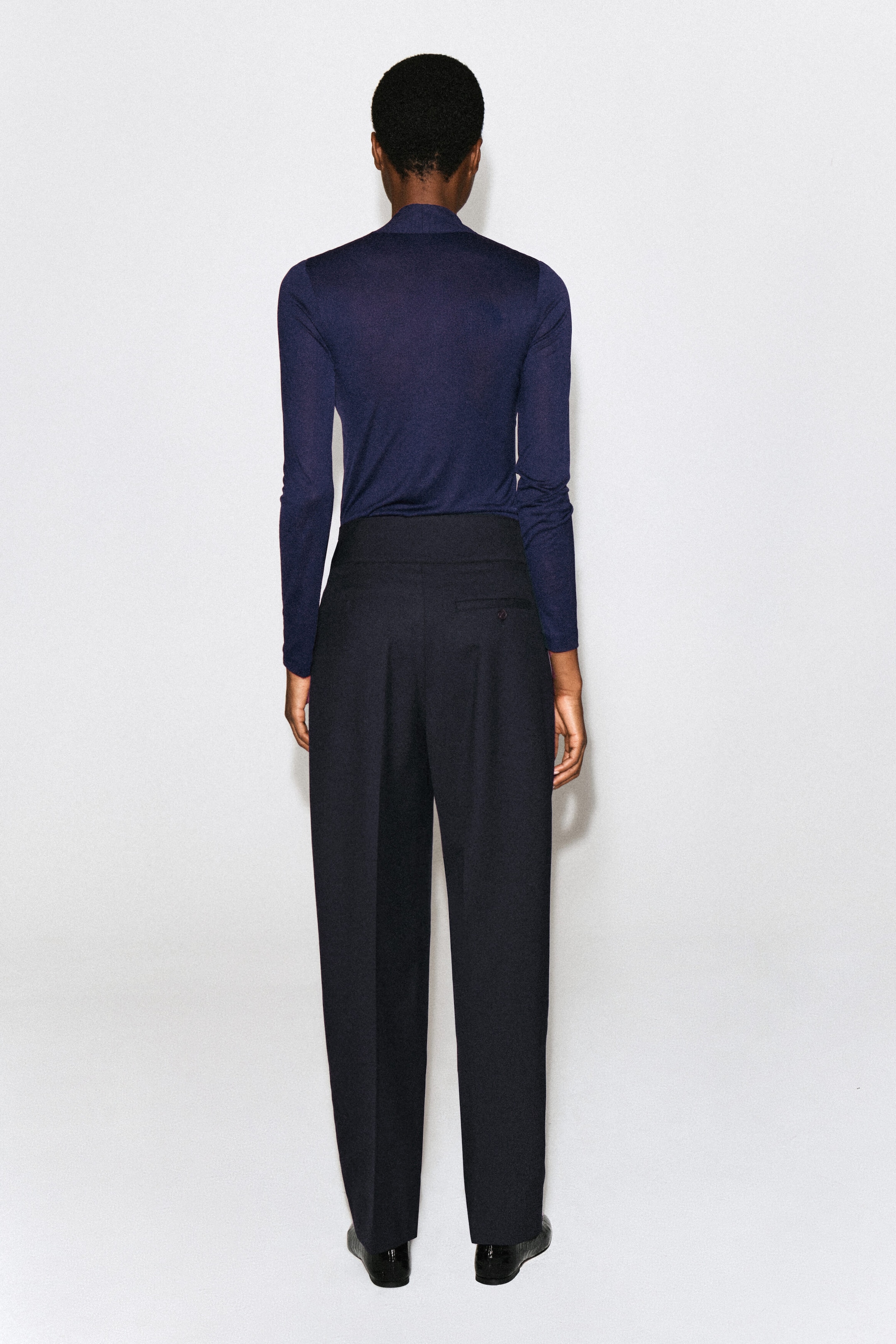 A dark blue, long-sleeved jersey top is worn by the model, shown from the back. The slim-fit top features a draped neckline at the nape and is styled tucked into dark, high-waisted trousers. A dark blue, long-sleeved jersey top is worn by the model, shown from the back. The slim-fit top features a draped neckline at the nape and is styled tucked into dark, high-waisted trousers.