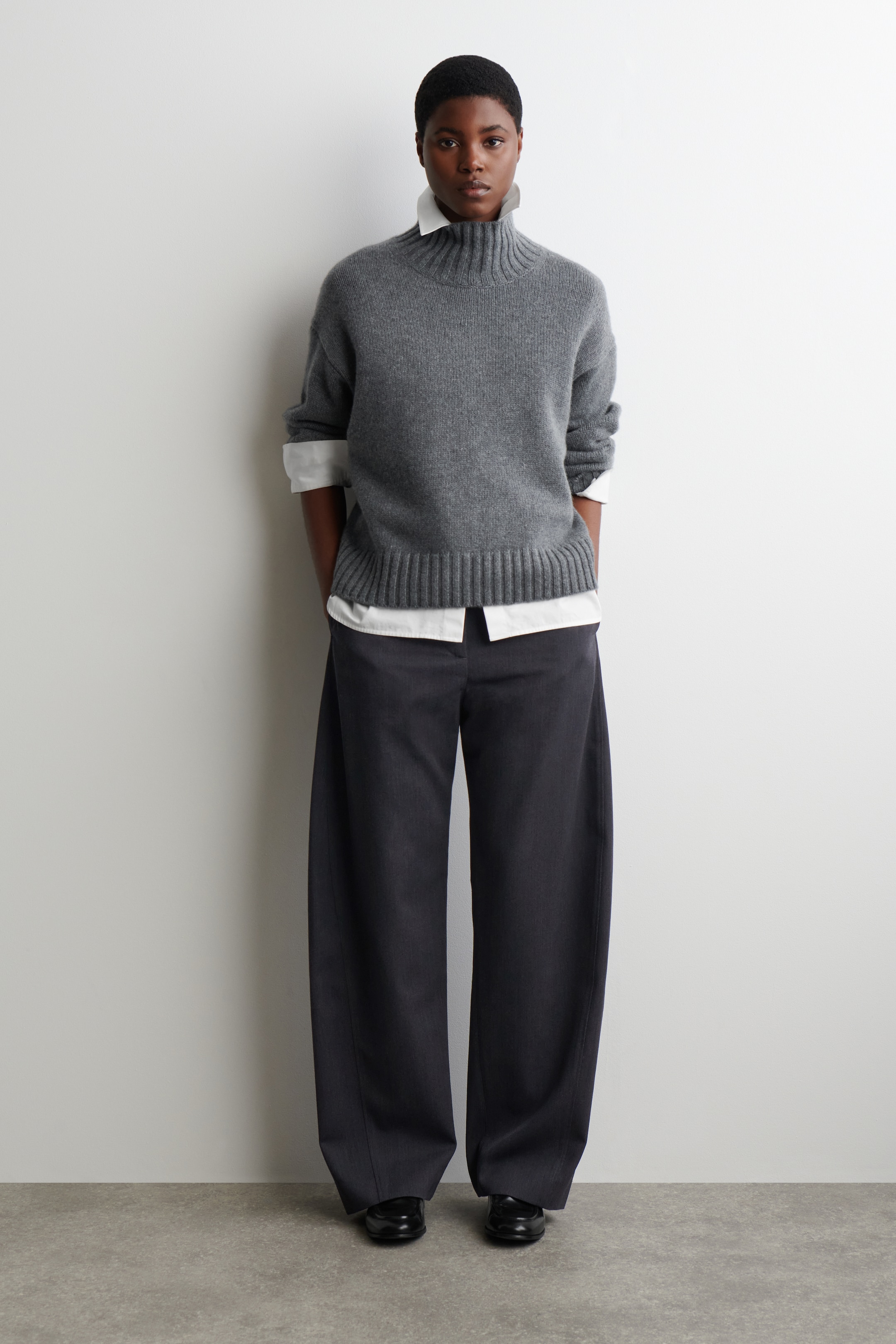 View larger image: A gray, front-facing, mock neck sweater is being displayed, featuring a ribbed collar and a wide ribbed hem with a relaxed fit. It is being layered over a white collared shirt, with the shirt's collar and cuffs visibly extending, and styled with dark wide-leg trousers and black flat shoes.