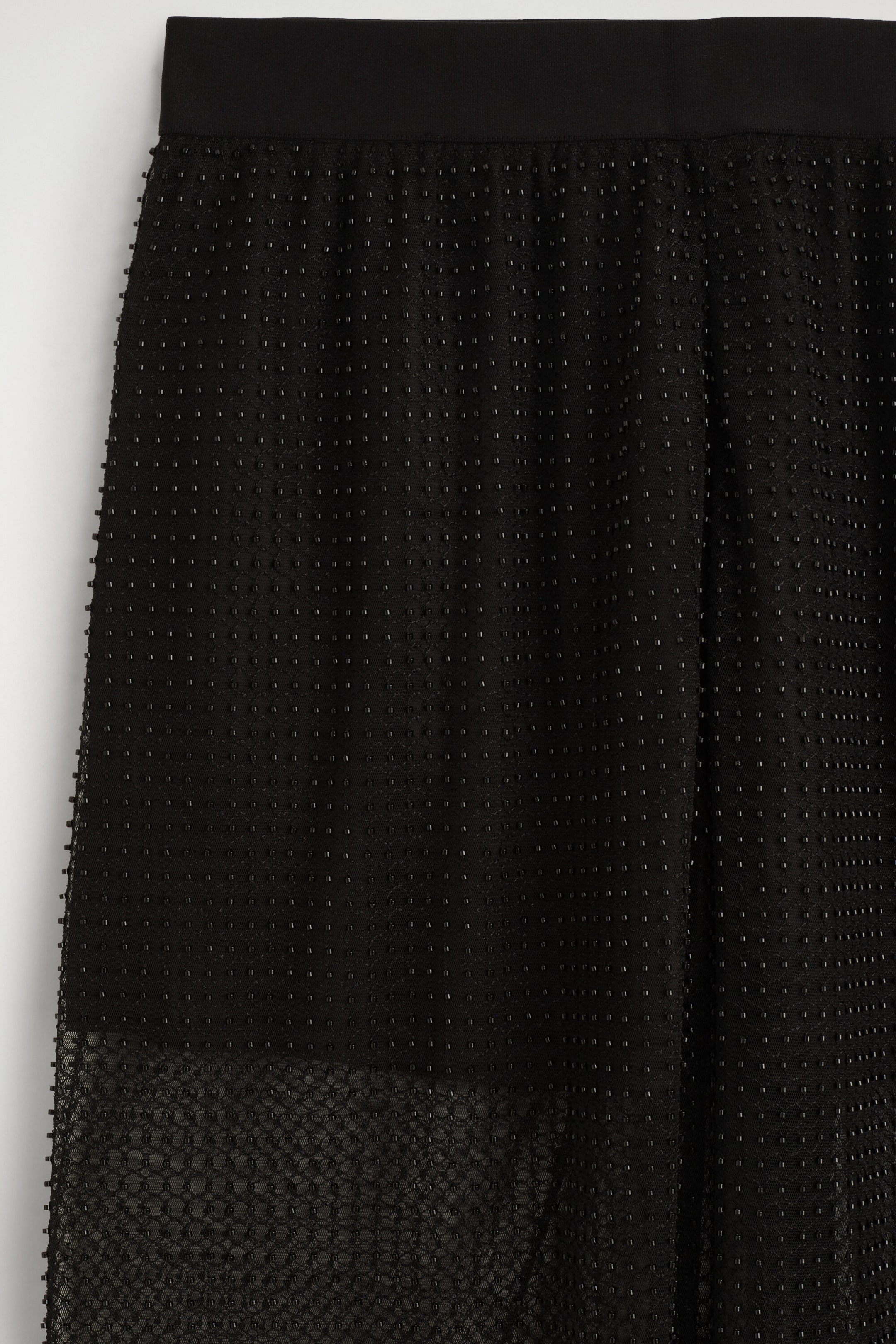 The black beaded knitted trousers feature a wide, smooth elastic waistband and a semi-sheer mesh body adorned with vertical rows of tiny black beads. A solid black lining covers the upper portion, transitioning into a sheer, patterned knit section at the hem.
