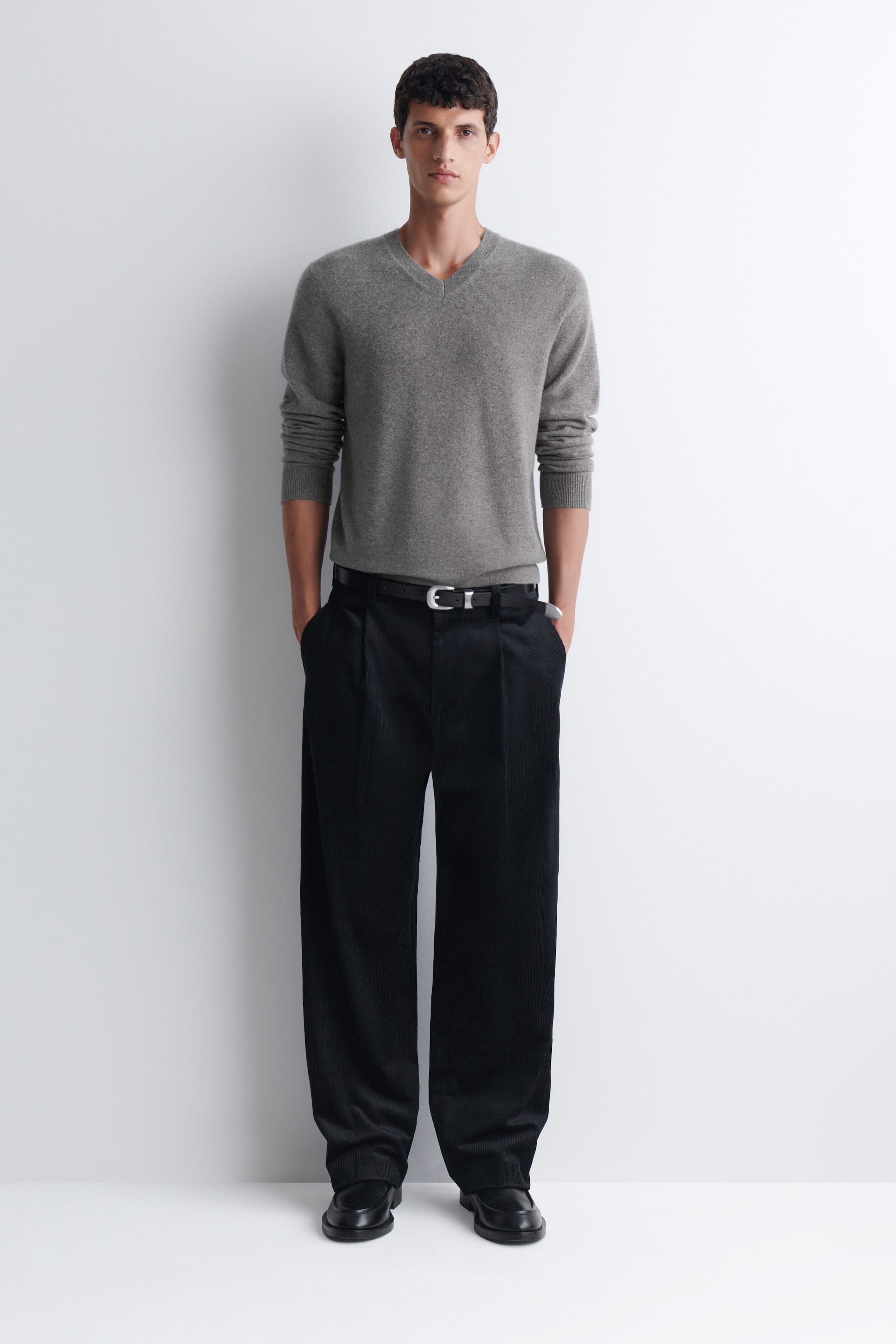 Pleated relaxed-fit tapered corduroy pants in black are worn by the model, with hands placed in the side pockets. The pants are styled with a tucked-in grey V-neck sweater, a black leather belt, and black dress shoes. Pleated relaxed-fit tapered corduroy pants in black are worn by the model, with hands placed in the side pockets. The pants are styled with a tucked-in grey V-neck sweater, a black leather belt, and black dress shoes.