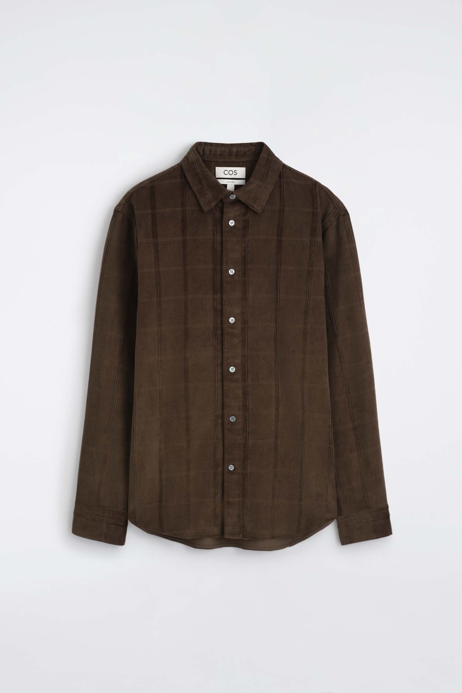 Una camicia in velluto a coste marrone scuro è mostrata frontalmente, con colletto a punta, chiusura frontale con bottoni e maniche lunghe con polsini. La camicia mette in evidenza un tessuto testurizzato con coste verticali in velluto e un delicato motivo a griglia.