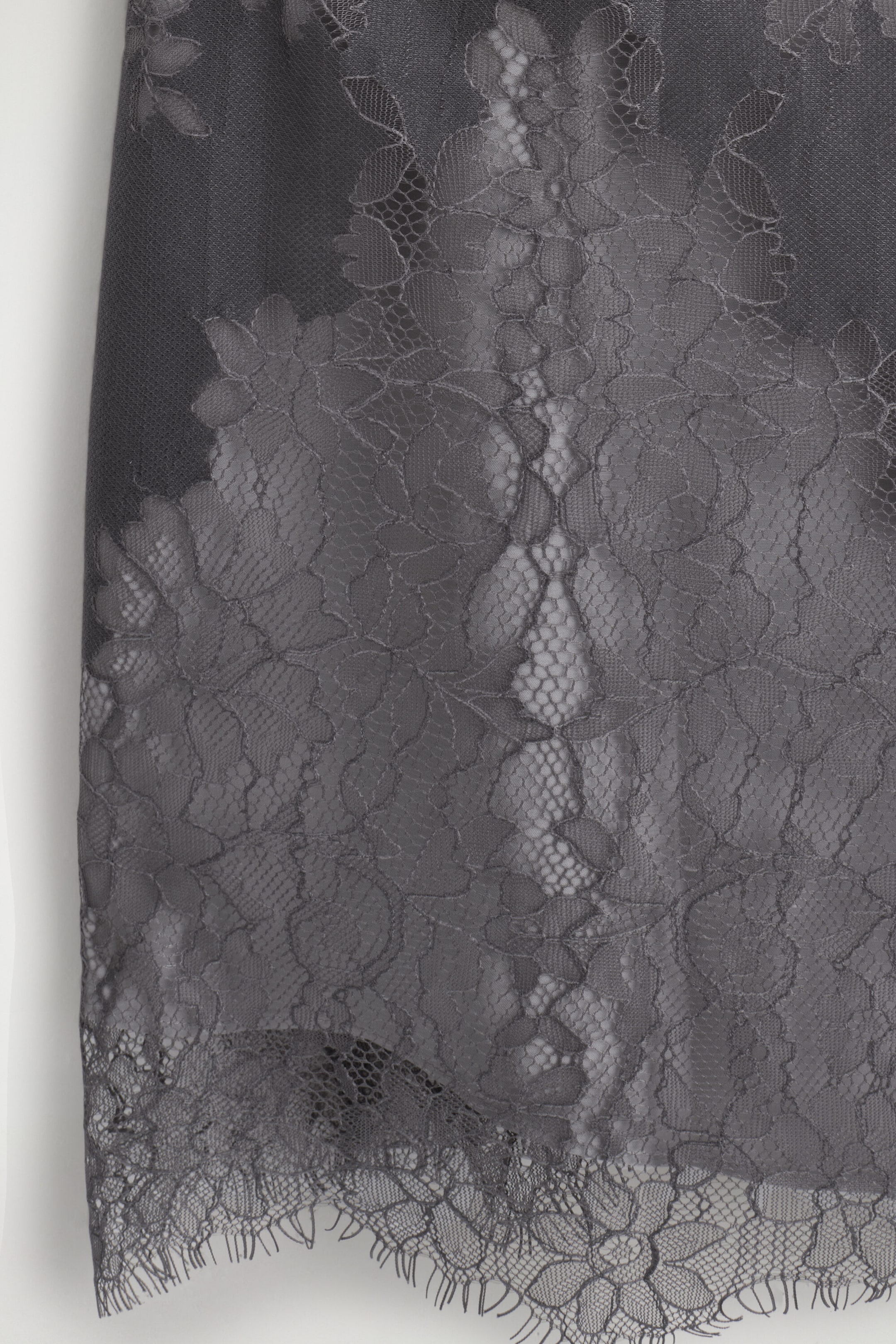 The LACE-OVERLAY CAMI TOP features an intricate dark grey lace overlay layered over a solid fabric base, highlighting a detailed floral pattern. The lace trim finishes with a delicate scalloped eyelash hem at the bottom. The LACE-OVERLAY CAMI TOP features an intricate dark grey lace overlay layered over a solid fabric base, highlighting a detailed floral pattern. The lace trim finishes with a delicate scalloped eyelash hem at the bottom.