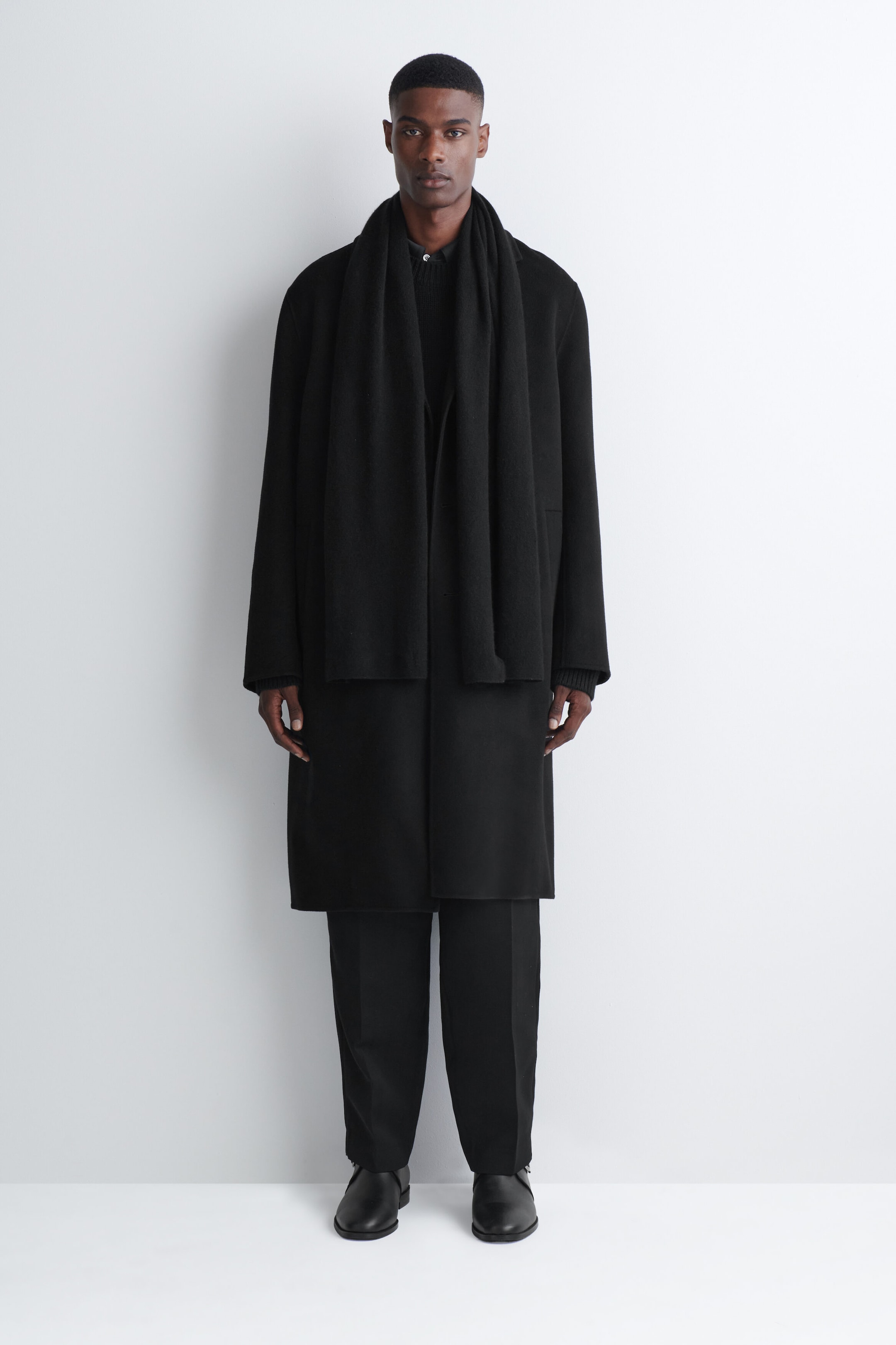 A black cashmere scarf is being worn draped around the model’s neck, with its long ends hanging straight down the front. The scarf is styled over a matching black overcoat as part of a layered, monochromatic outfit.
